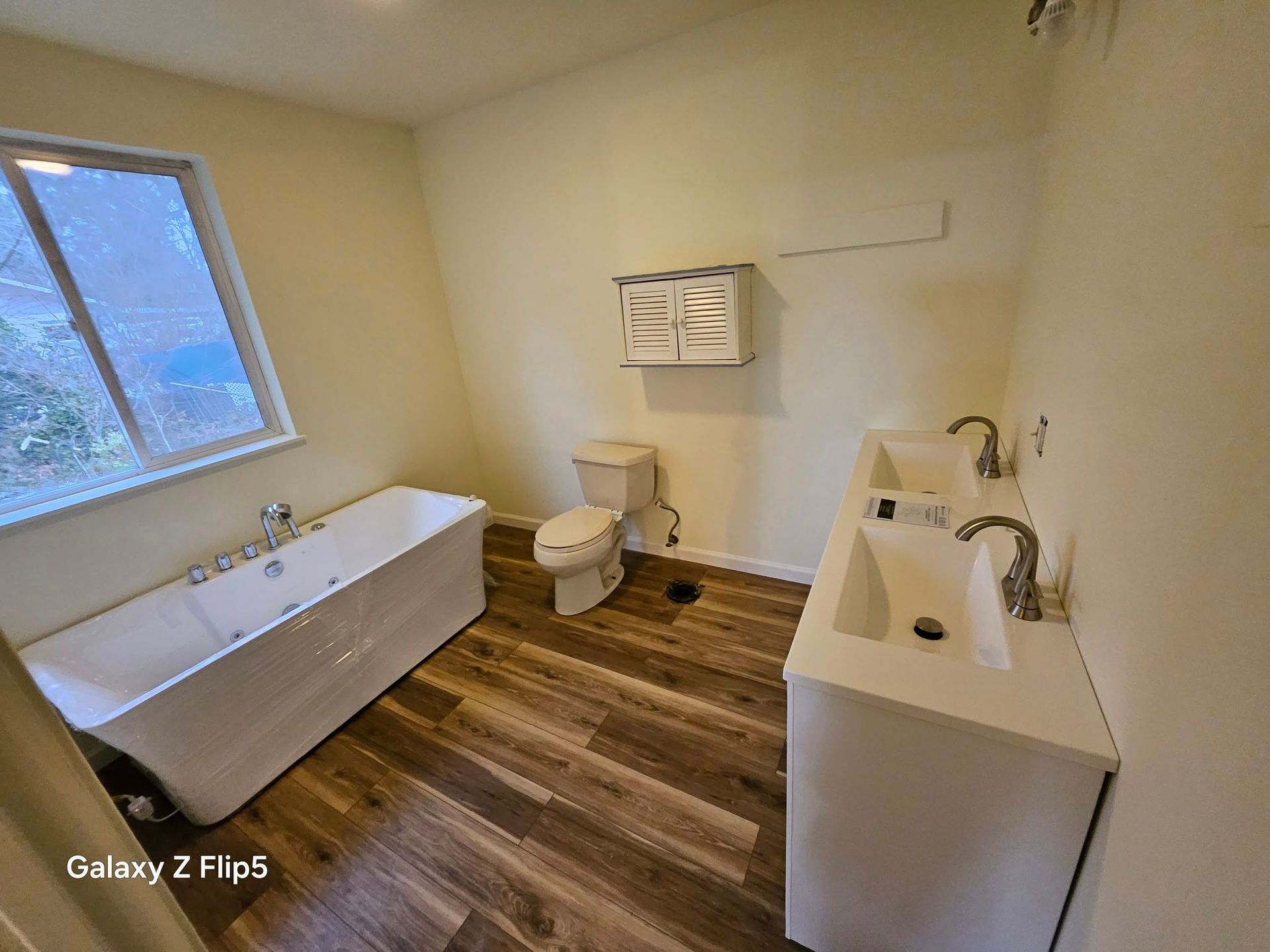 Bathroom with a freestanding tub, dual sinks, toilet, and window. Brown wood-look flooring, cream walls.
