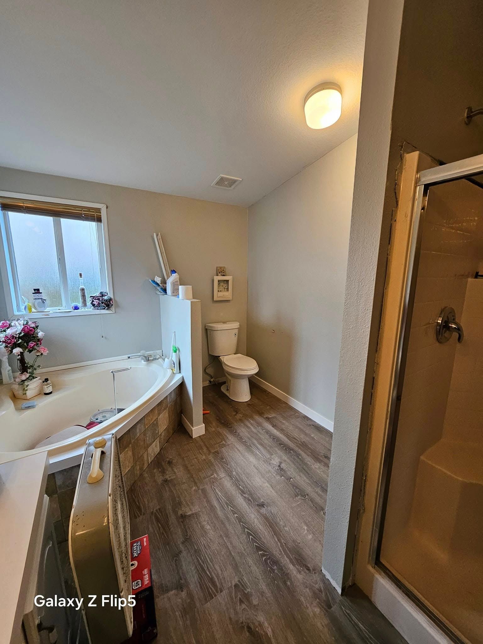 Bathroom with a tub, toilet, and shower. Features gray walls, wood-look floor, and a window.