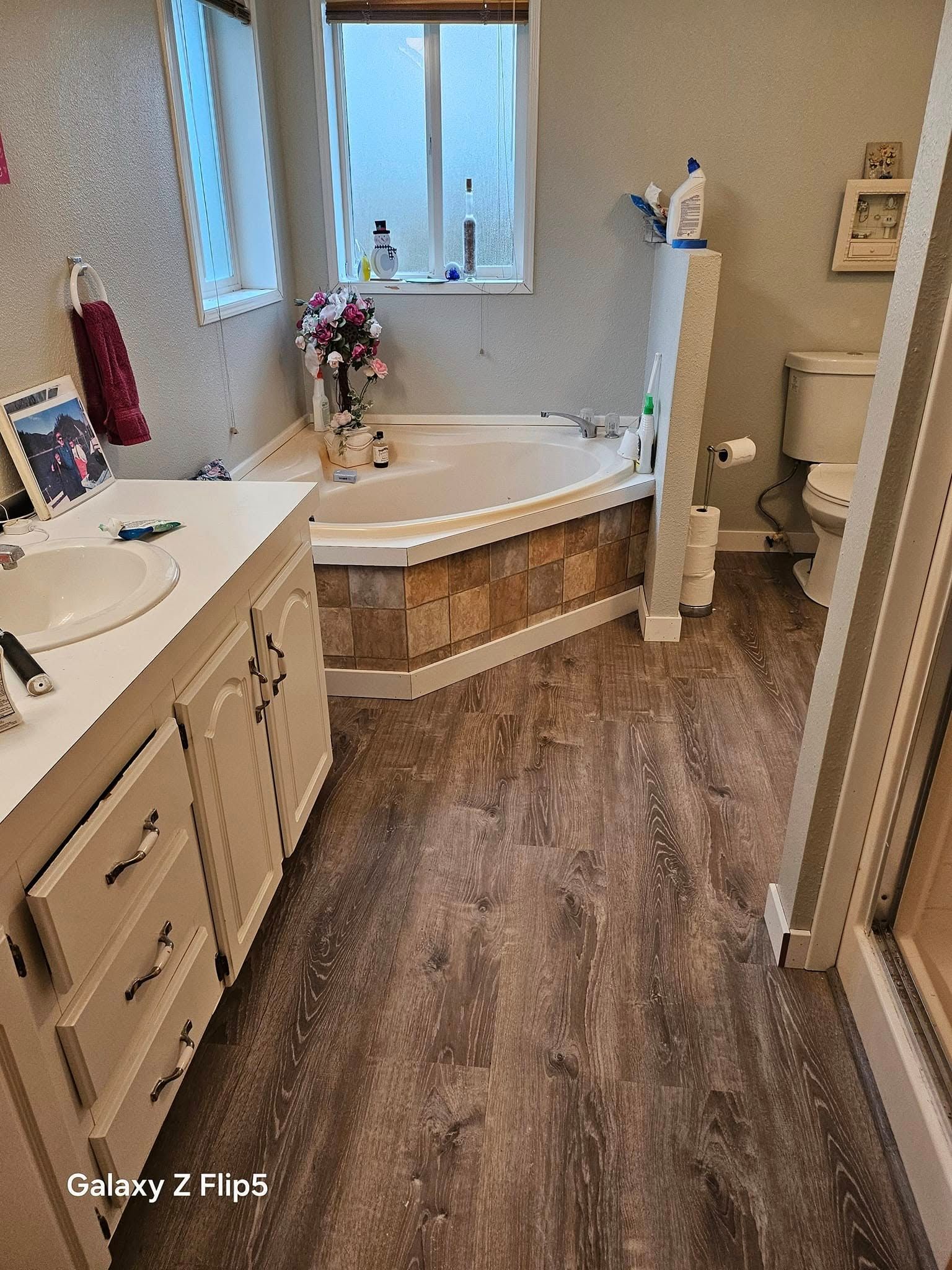 Bathroom with wooden floor, white vanity, corner tub, and a toilet near a window.