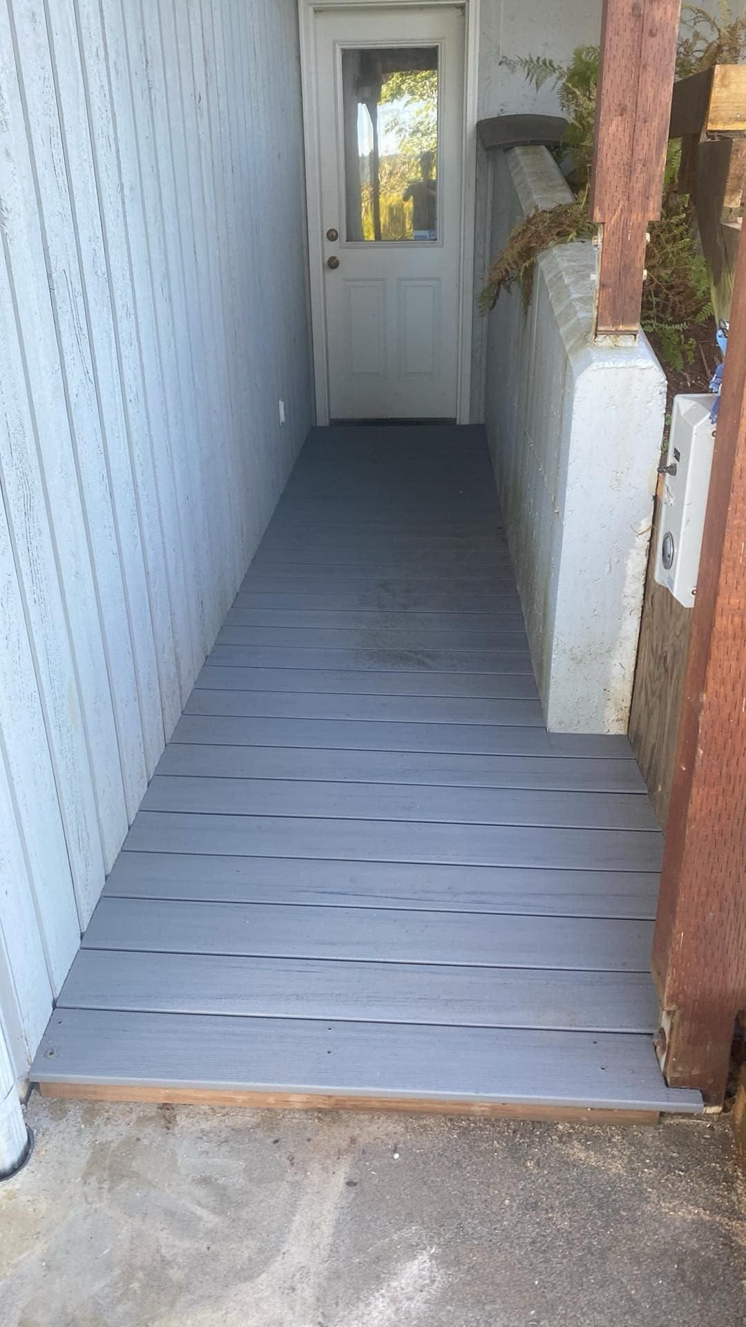 A gray wooden walkway leading to a white door.  A white wall and a concrete pillar are on the sides.