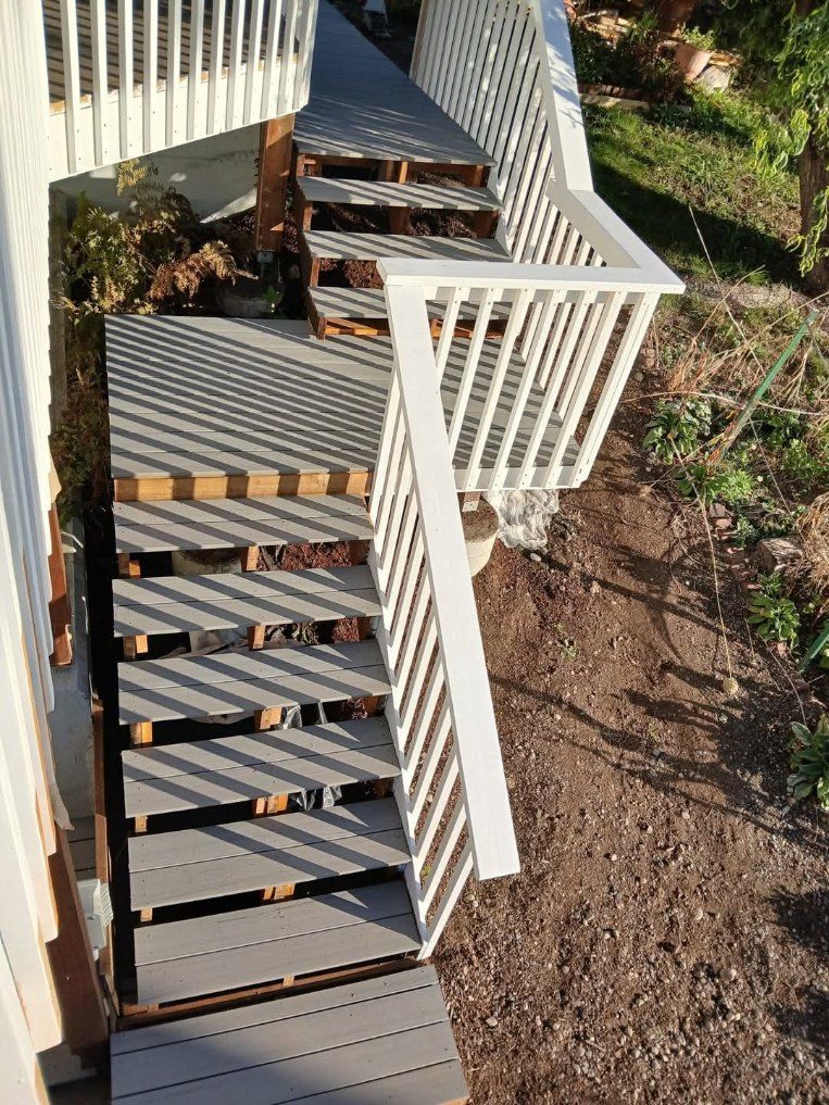 Wooden outdoor stairs with white railing lead down from a deck to a dirt yard.