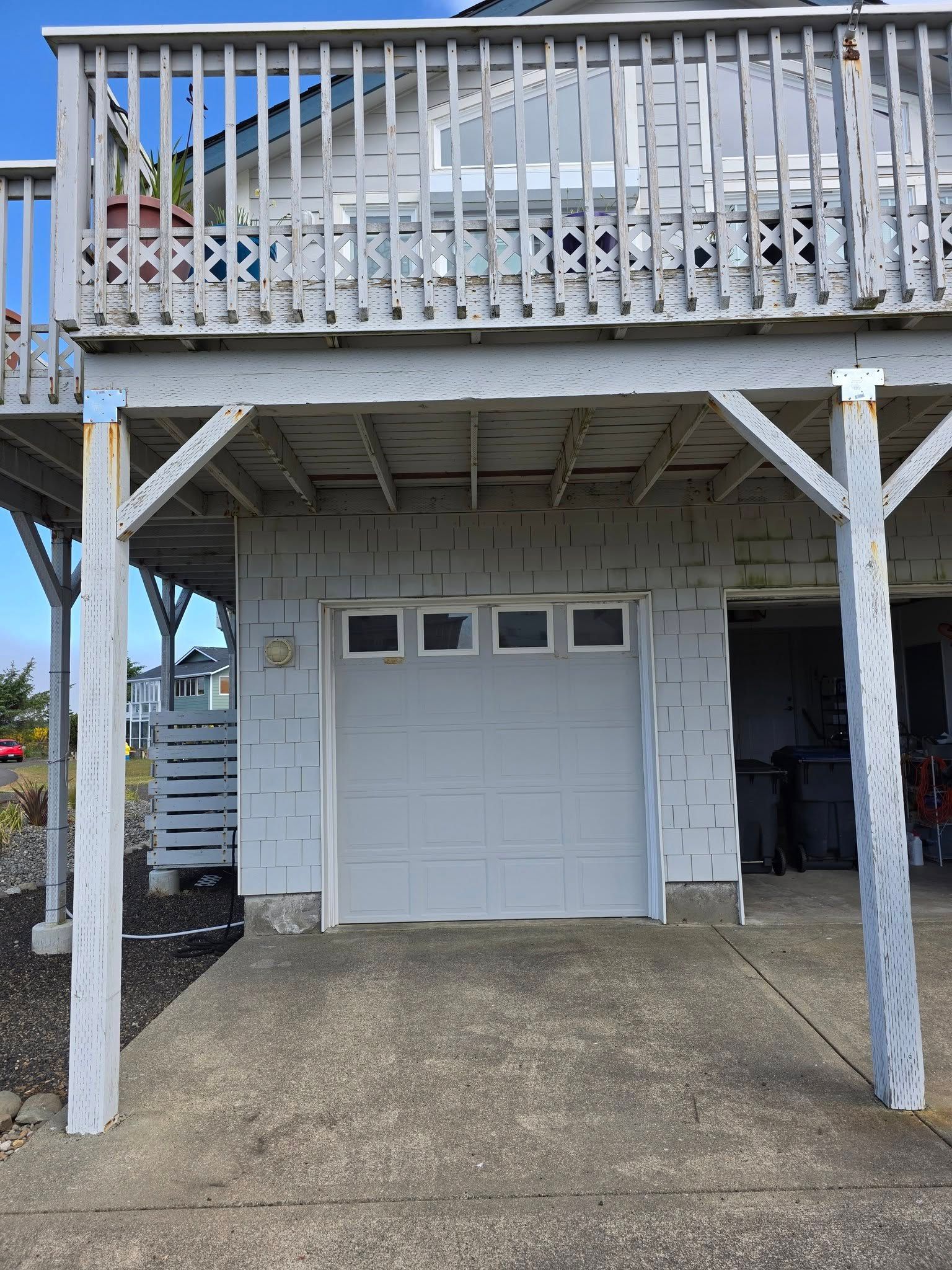 Garage under a raised house with a deck; gray siding and garage door. Concrete driveway.