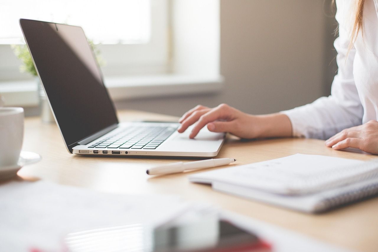 Photo of a woman working on her desk, with her laptop, a notebook and a pen