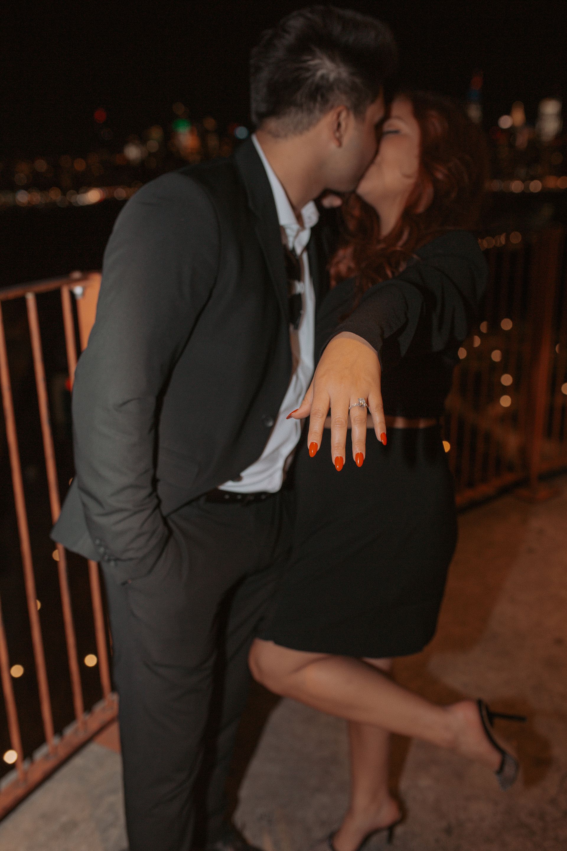 A man is kissing a woman on the cheek while she shows off her engagement ring.