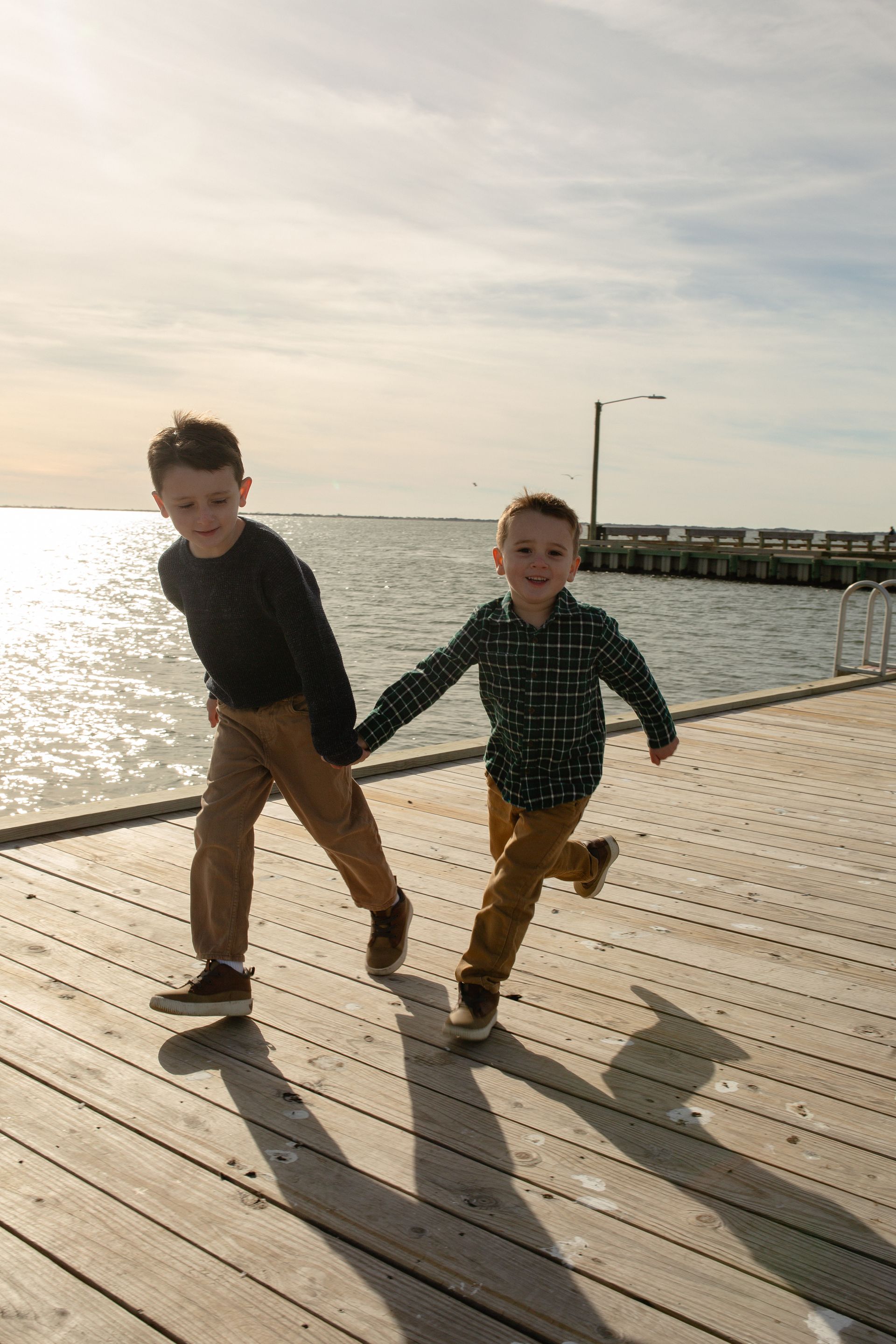 Two young boys are running on a wooden pier holding hands.