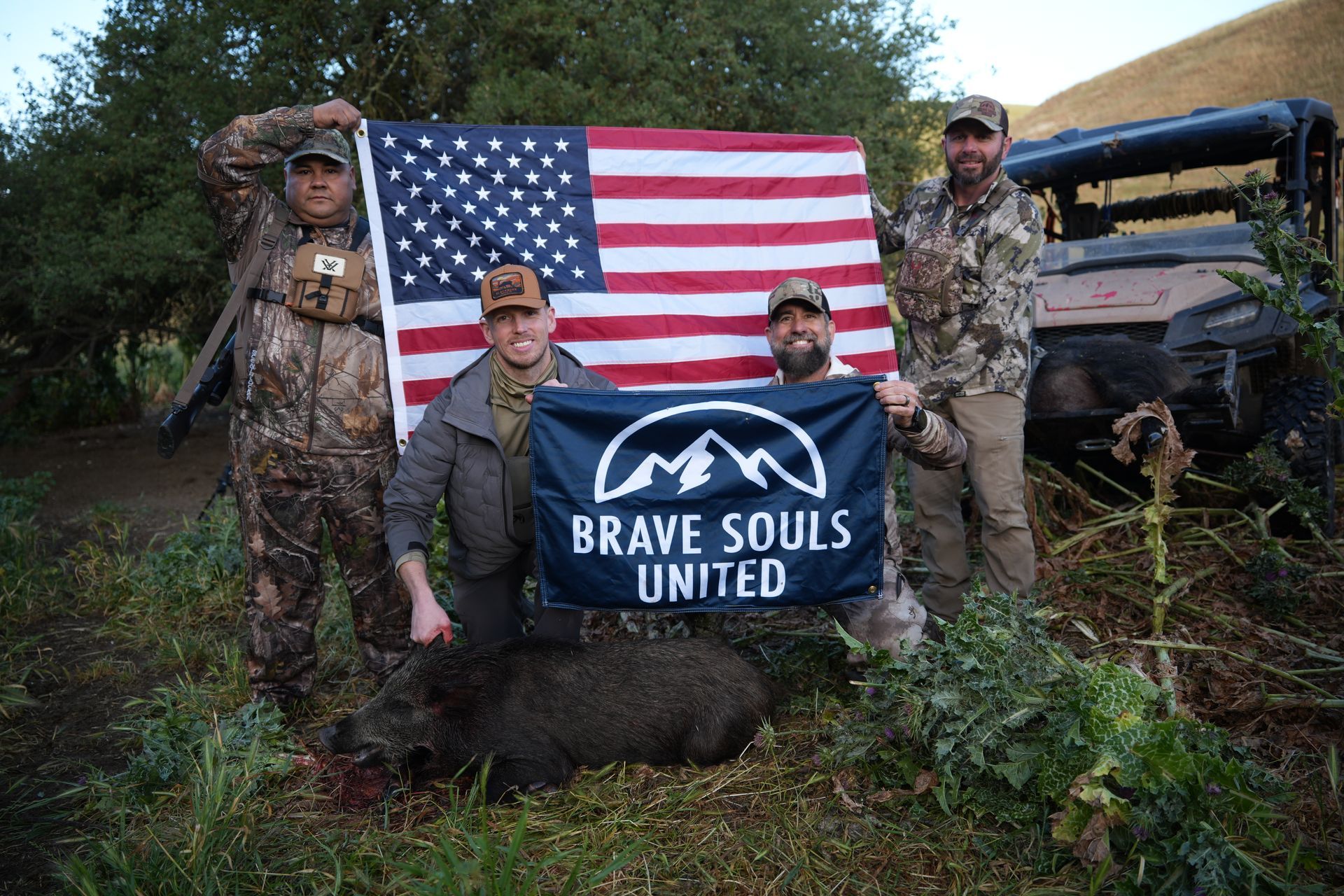 Three people in camouflage pose with an American flag and a “Brave Souls United” banner outdoors.