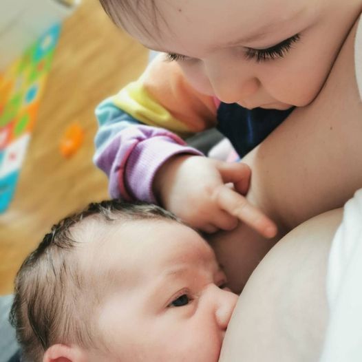 A baby is breastfeeding while another child looks on