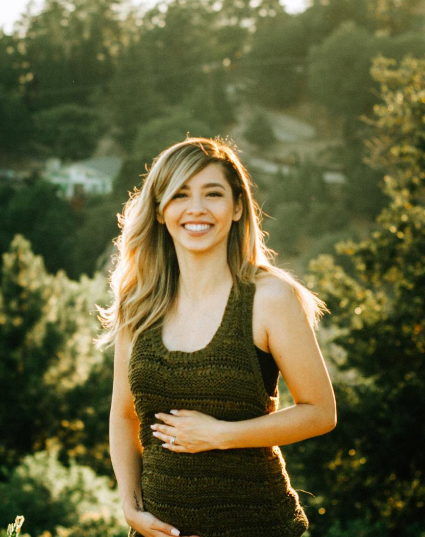 A pregnant woman in a green tank top is smiling and holding her belly.