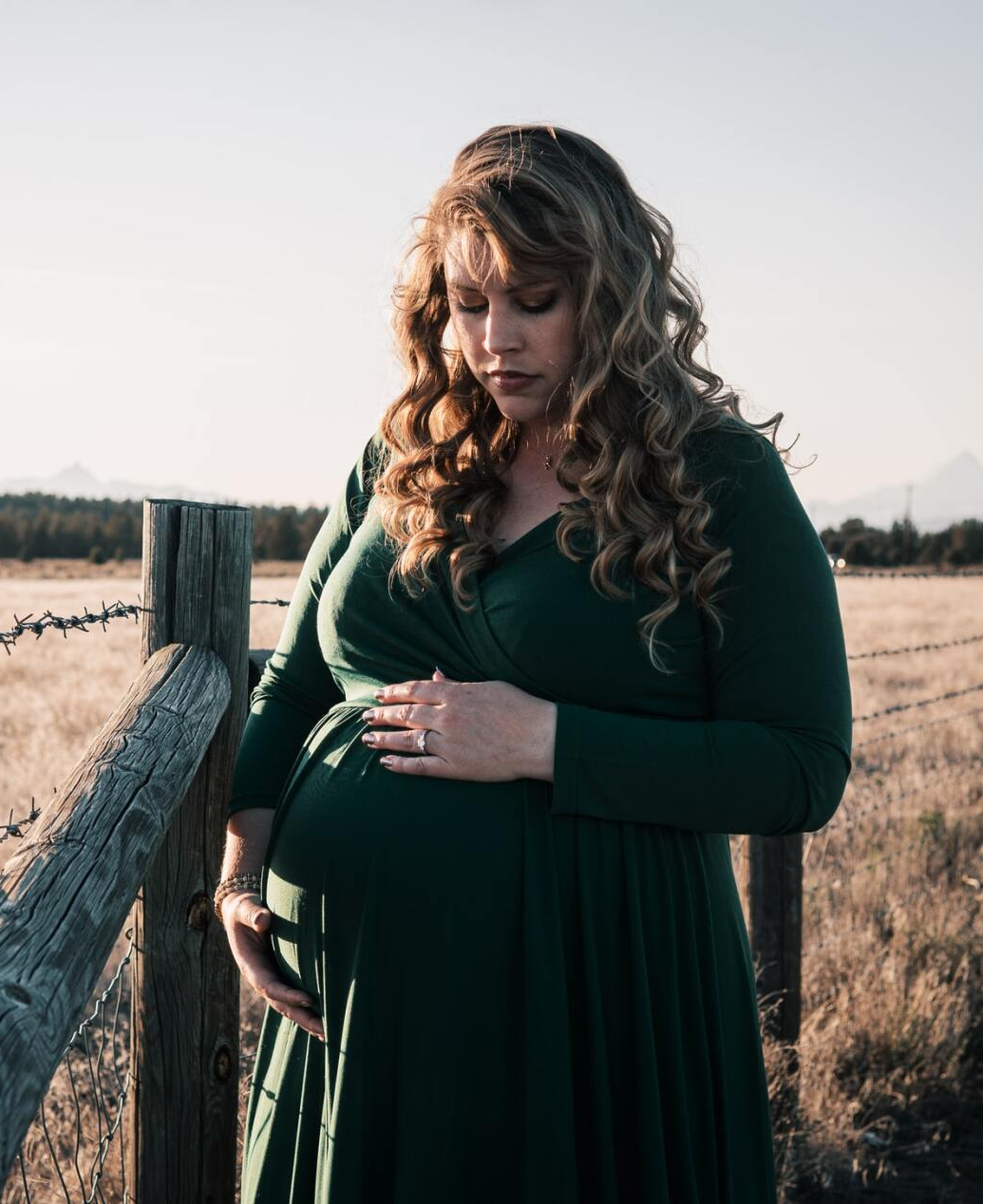 A pregnant woman in a green dress is standing next to a wooden fence holding her belly.