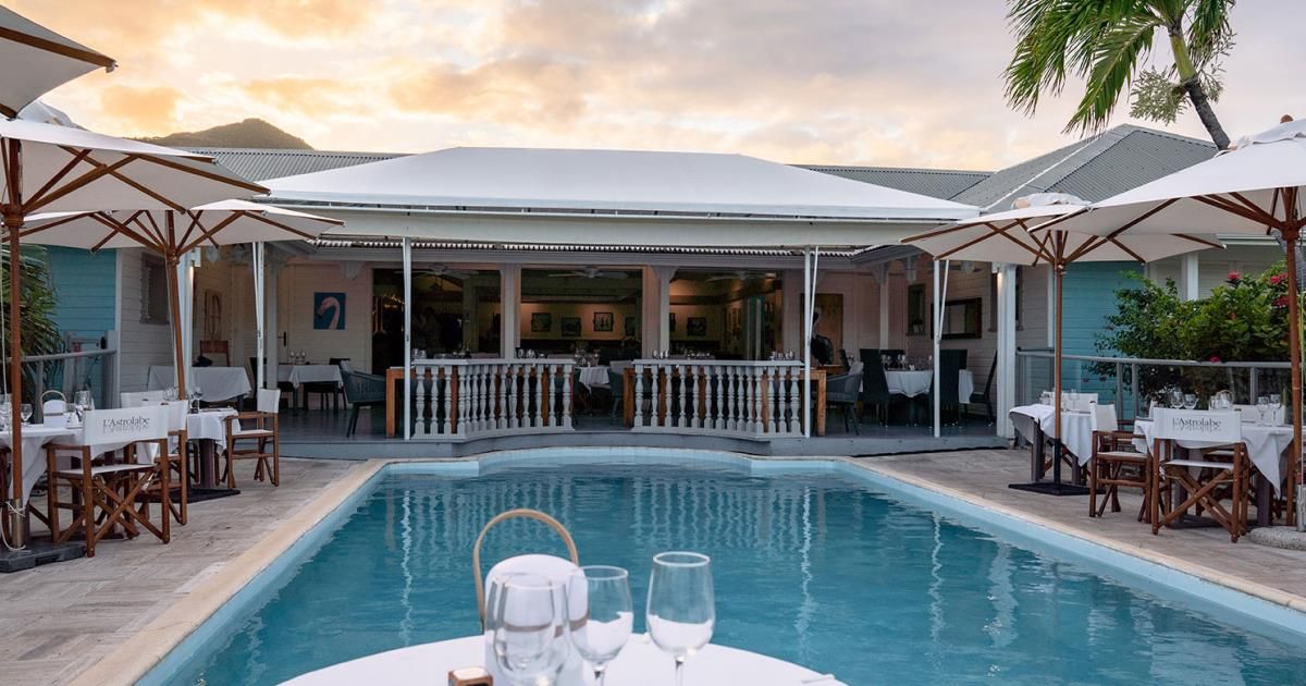 A swimming pool with tables and umbrellas in front of a restaurant.