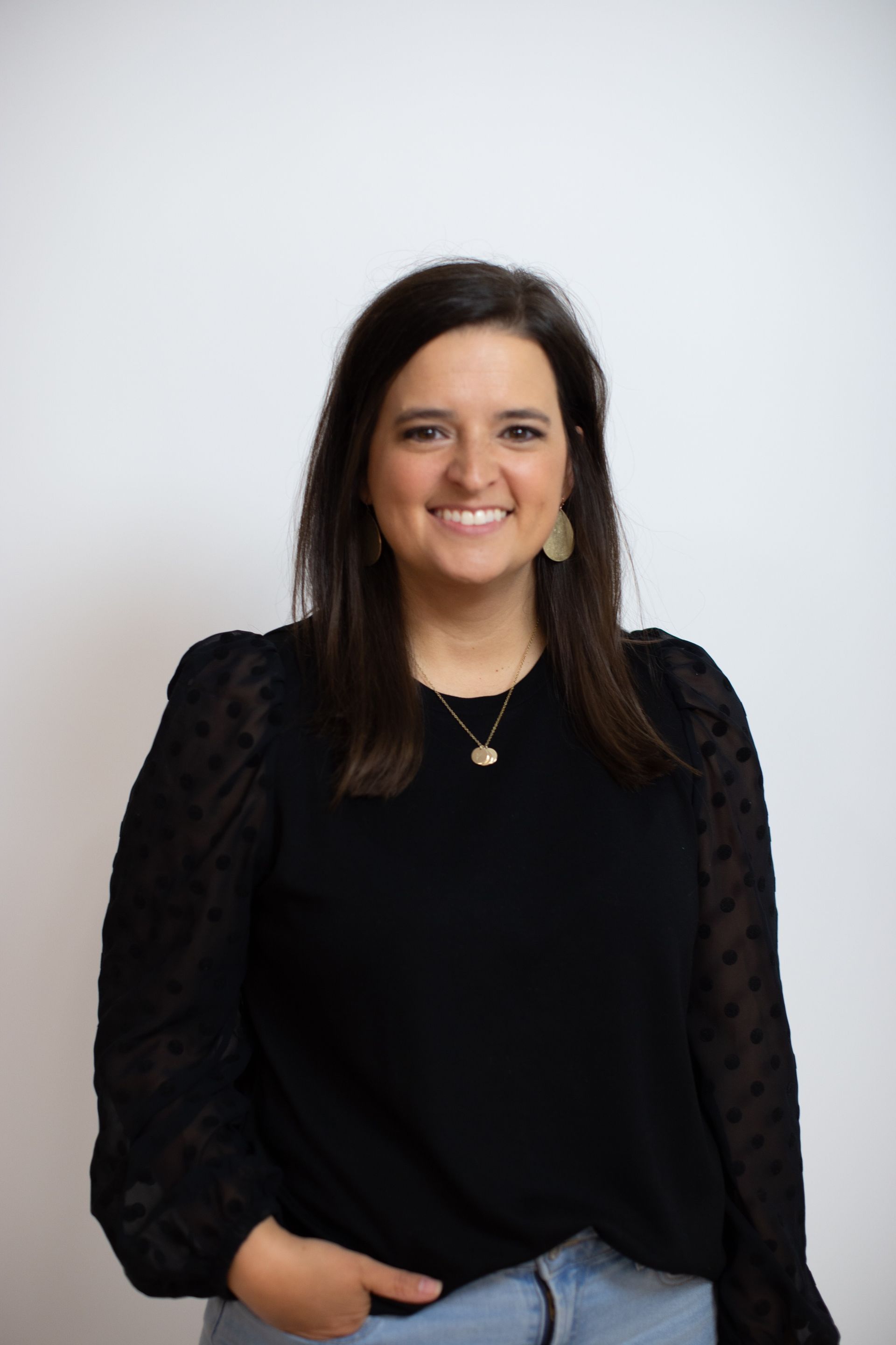 A smiling woman with dark hair wearing a black blouse and gold necklace against a white background. She has one hand in her pocket.
