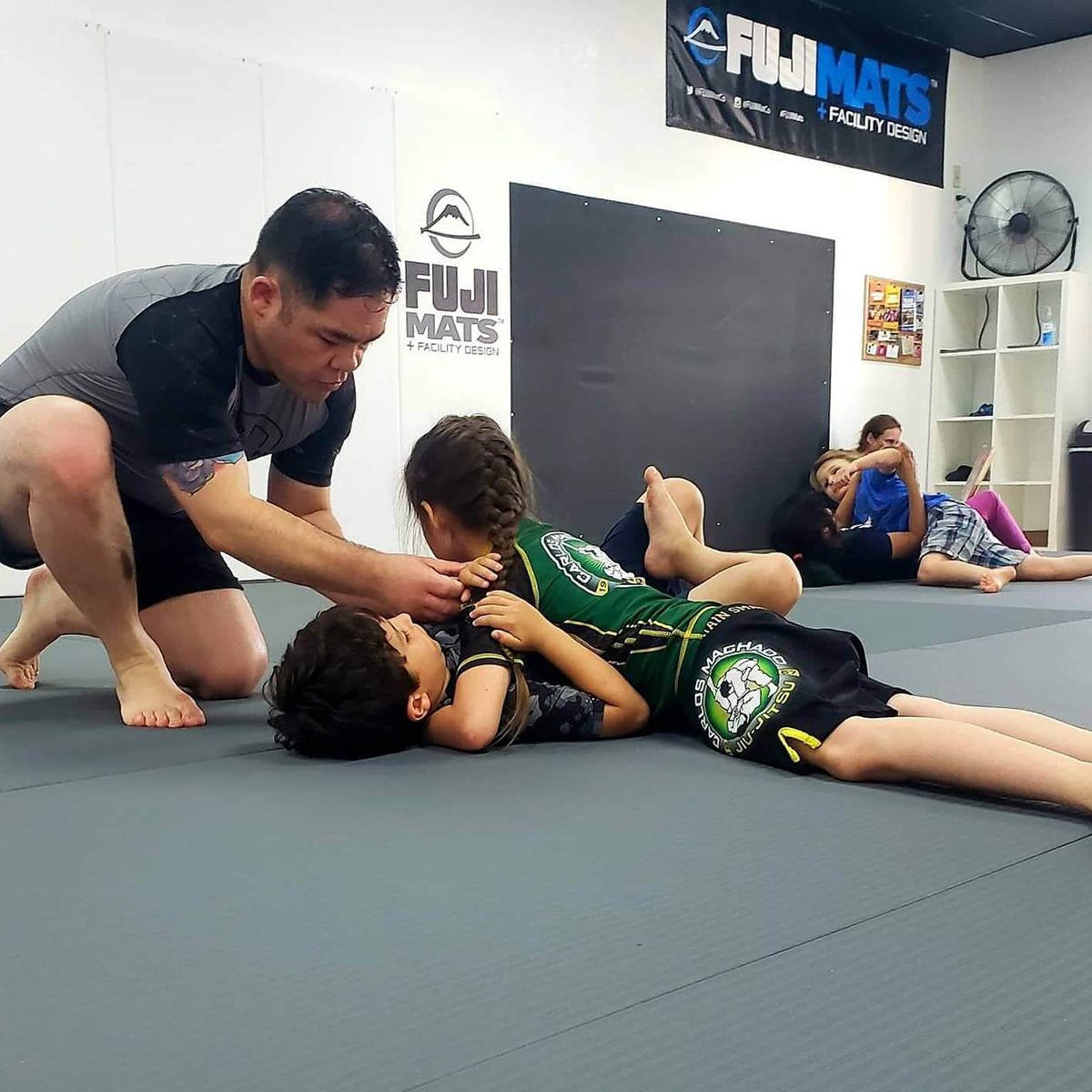 A man is kneeling down next to a child on a mat in a gym.