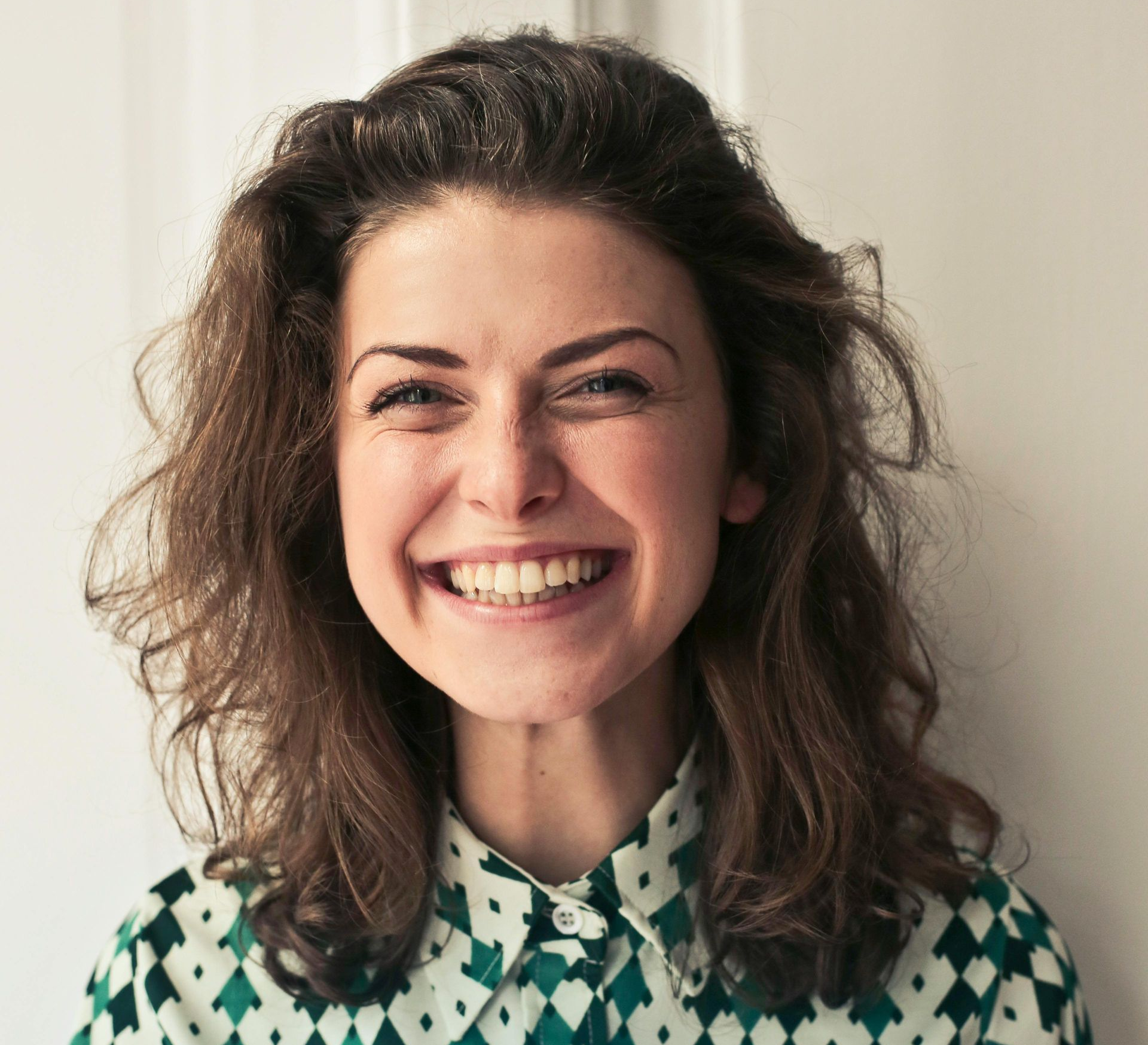 Woman with curly brown hair smiles widely, wearing a patterned shirt in a white setting.