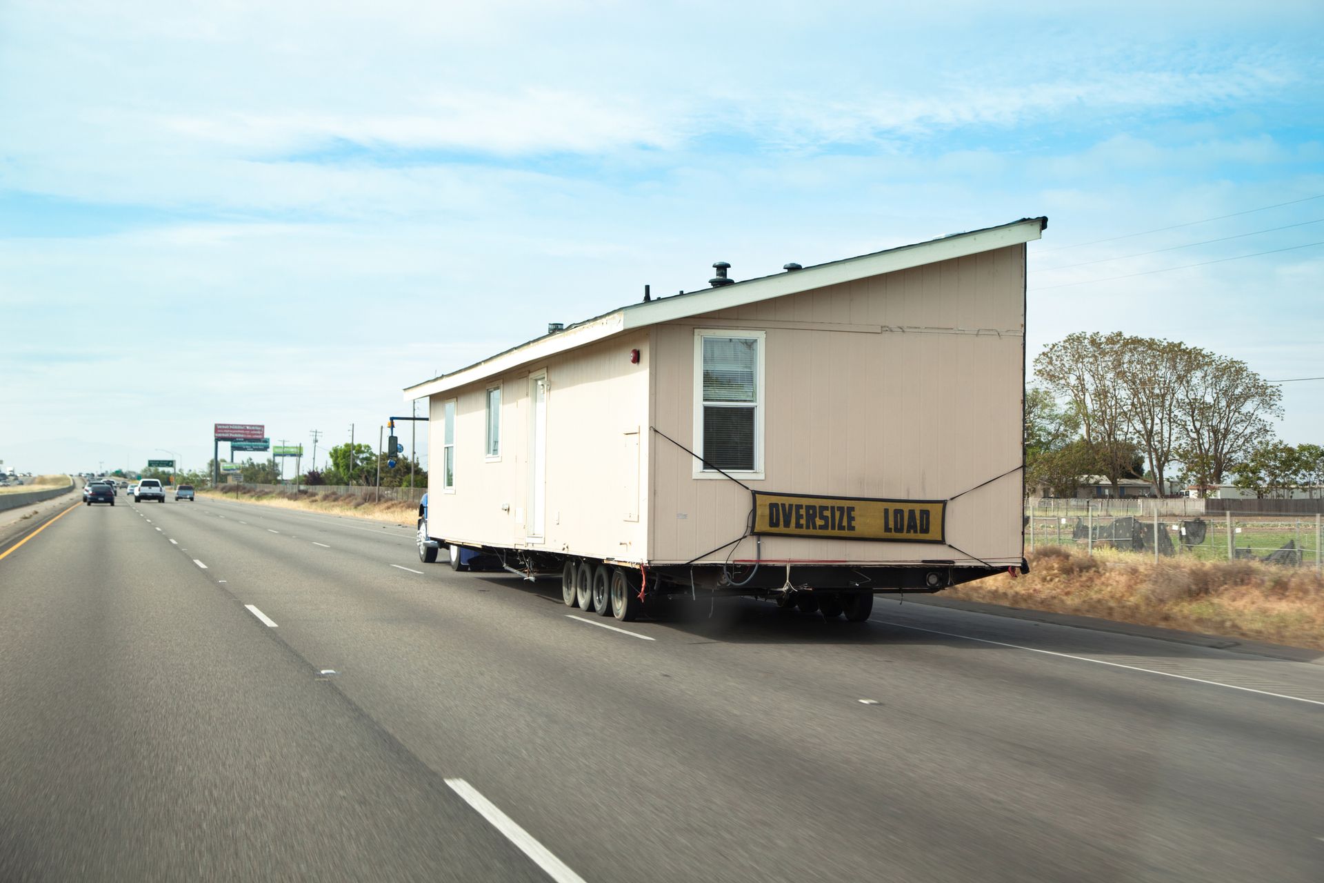 Mobile home being transported on a highway, light-colored with black tie-downs, under a blue sky.