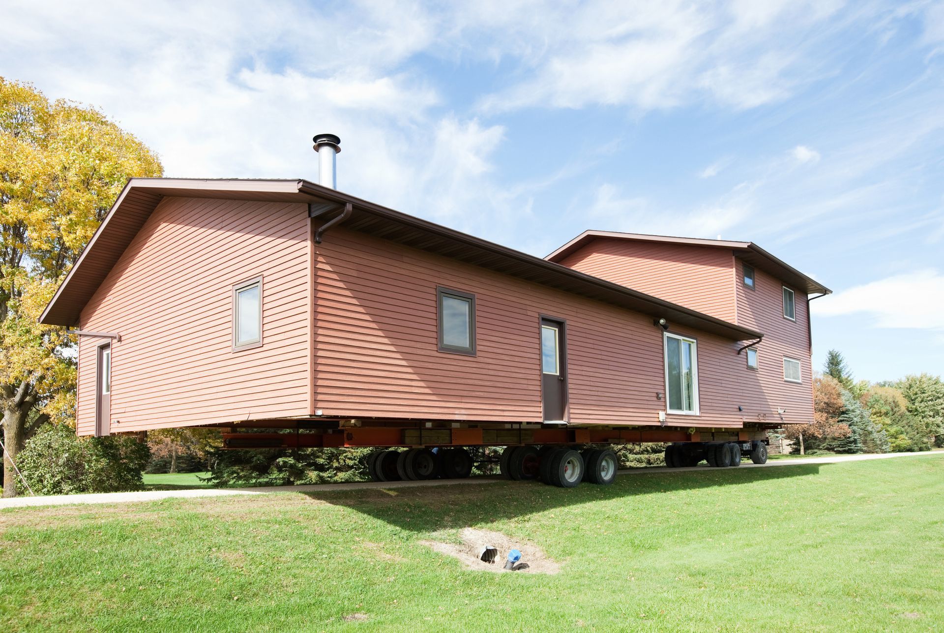 Brown house on a trailer on a green lawn under a blue sky with trees.