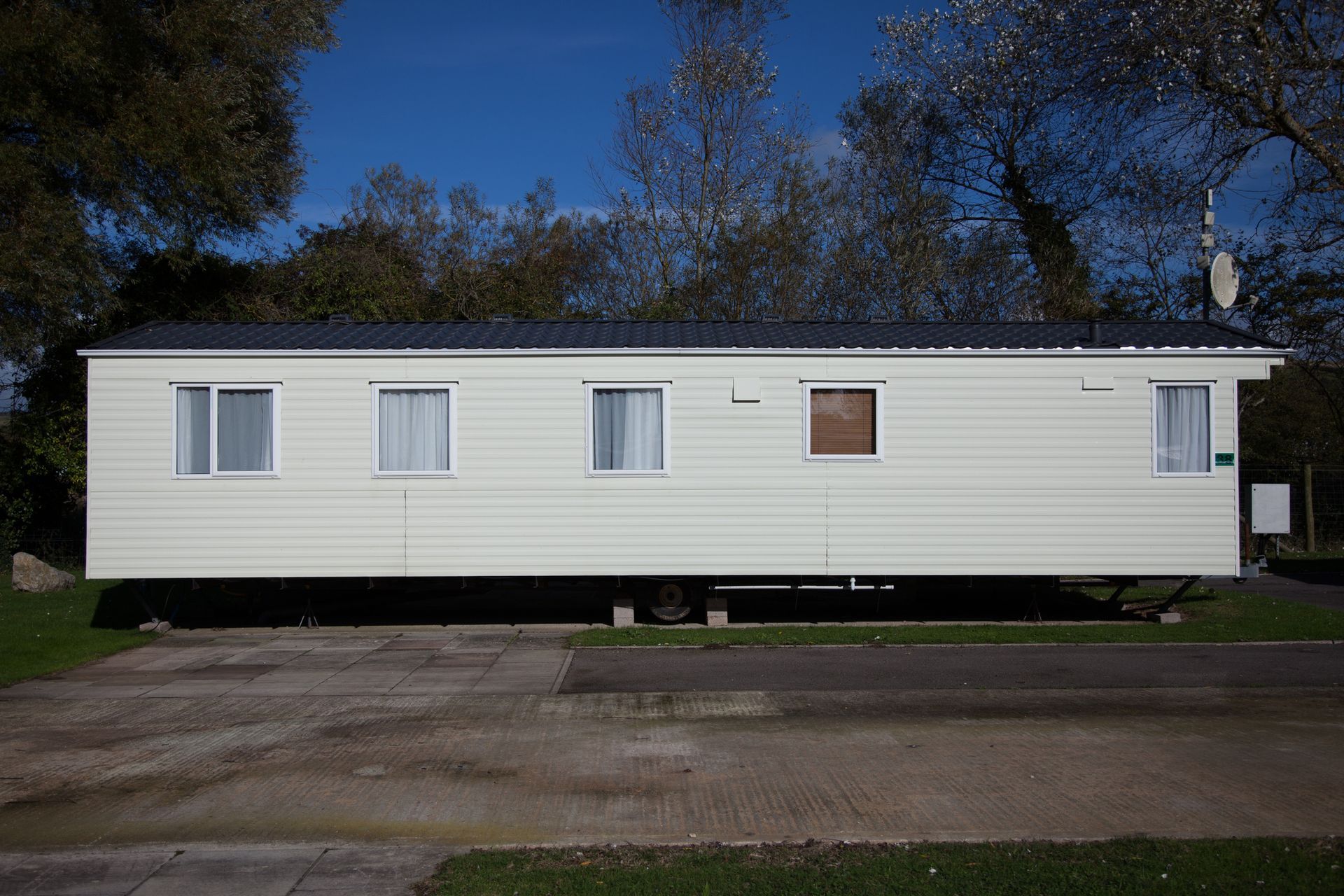 White mobile home on a gravel lot with blue sky and trees in the background.