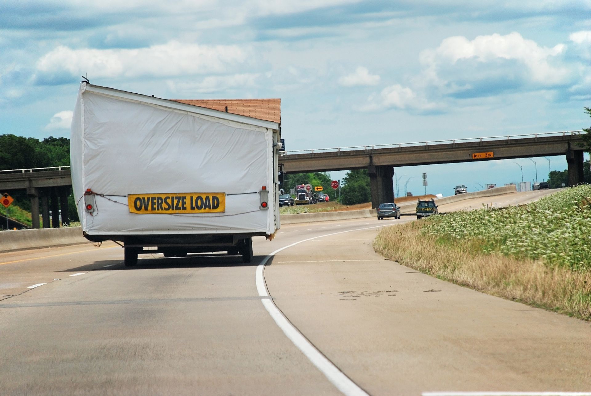 Truck labeled OVERSIZE LOAD driving on highway, approaching a low overpass.