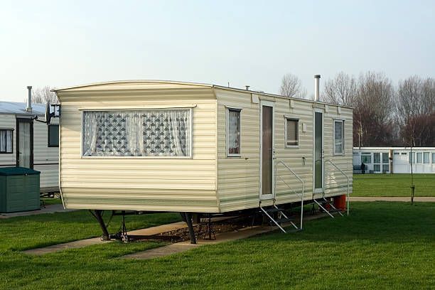 Tan mobile home on green grass, in a park. Blue sky, other homes in background.