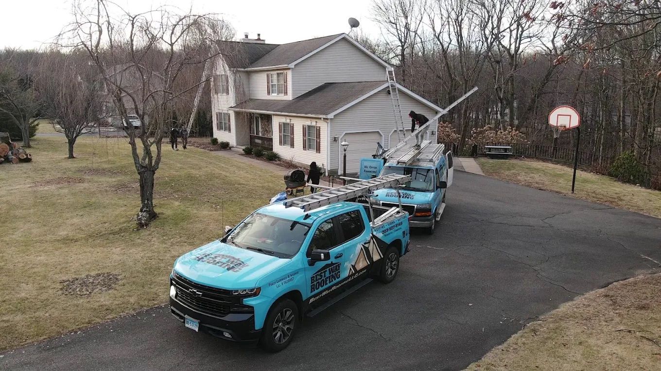 Blue truck with a ladder and aerial lift in front of a house. Men work on the roof.