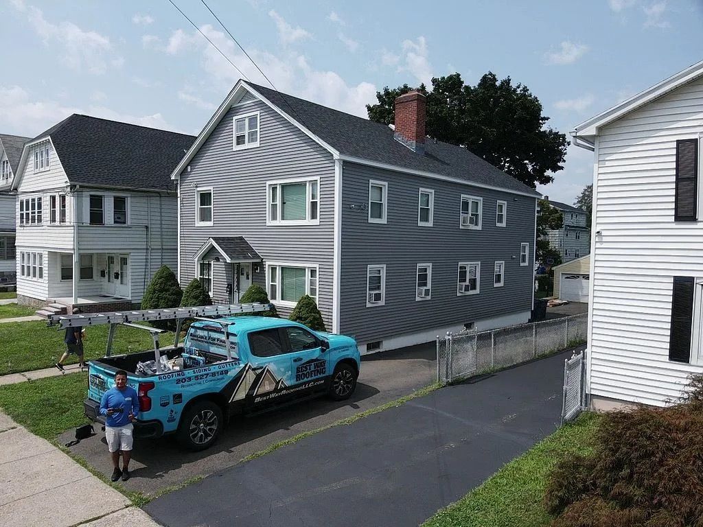 Blue truck with logo parked in front of gray house, man standing nearby. Other houses visible.
