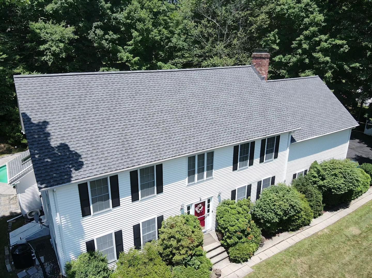 White two-story house with dark gray roof, black shutters, red door, and green bushes, surrounded by trees.