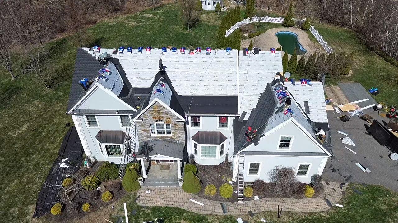 Aerial view of a house undergoing roof replacement with workers, white tarps, and a pool in the background.
