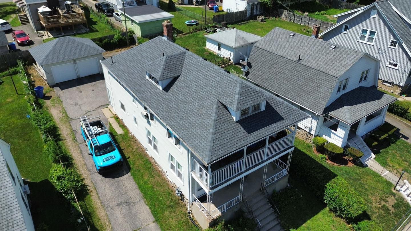 Aerial view of a gray-roofed house with a porch, surrounded by other houses and green lawns. A blue truck is parked nearby.