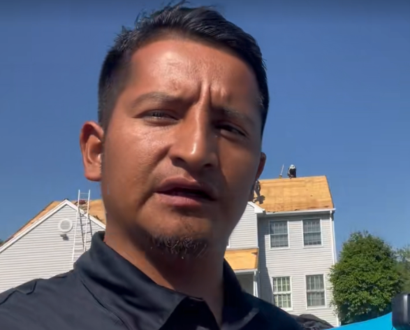 Man speaking in front of a house with roofers working; sunny day.