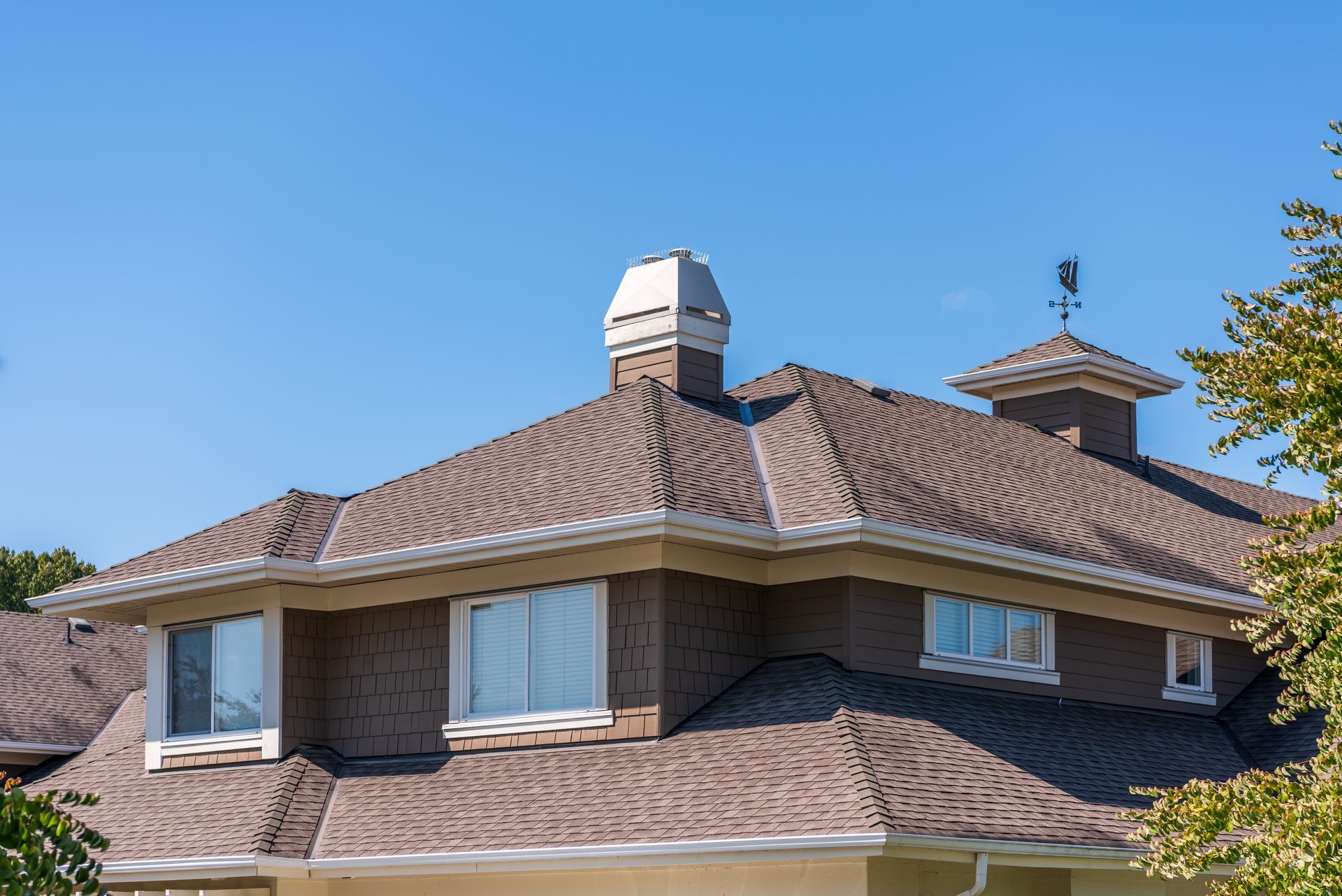 Brick house roof with brown shingles and white vent under a clear blue sky