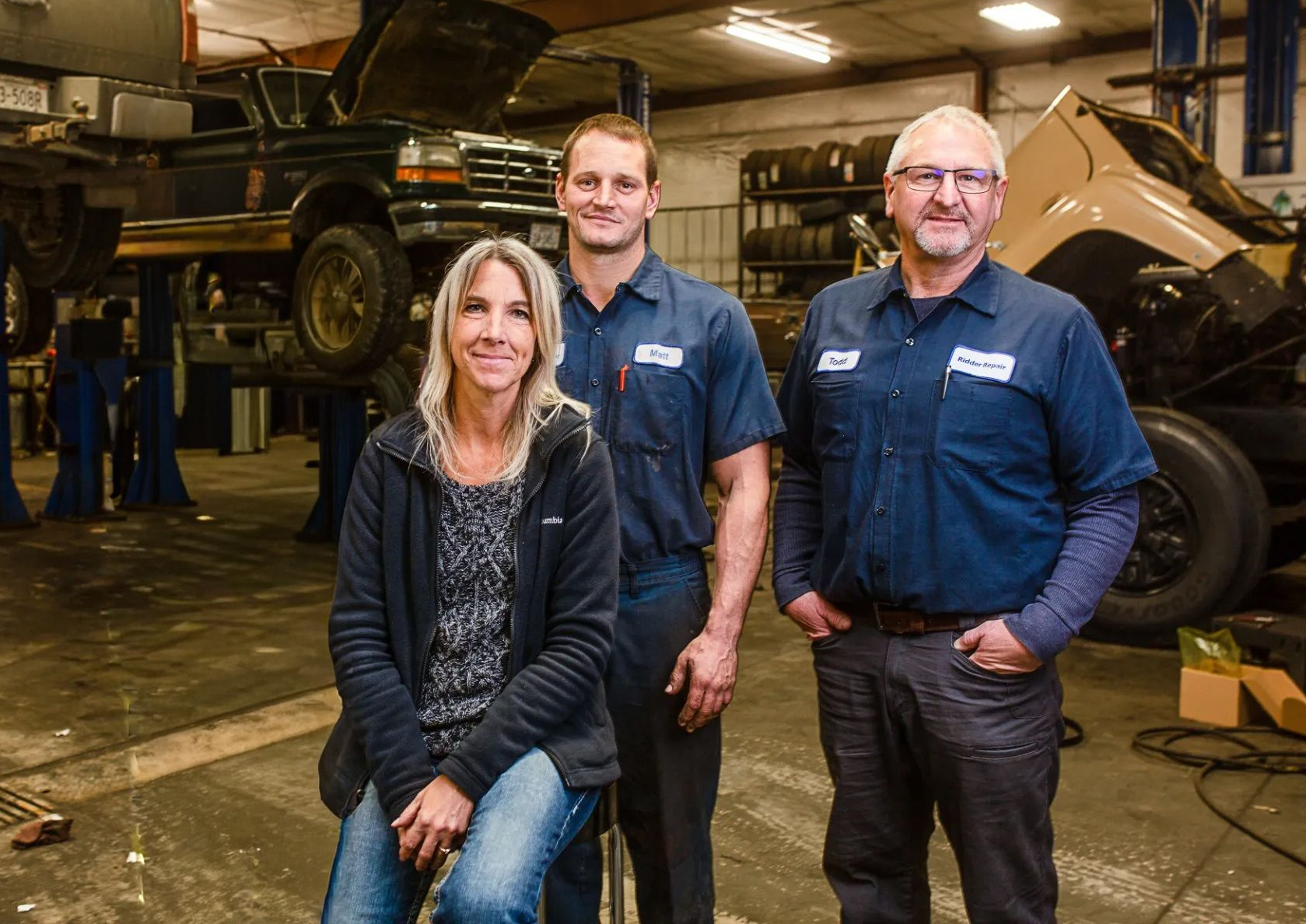 Three auto mechanics posing in a garage; woman seated, two men standing, vehicles in background.