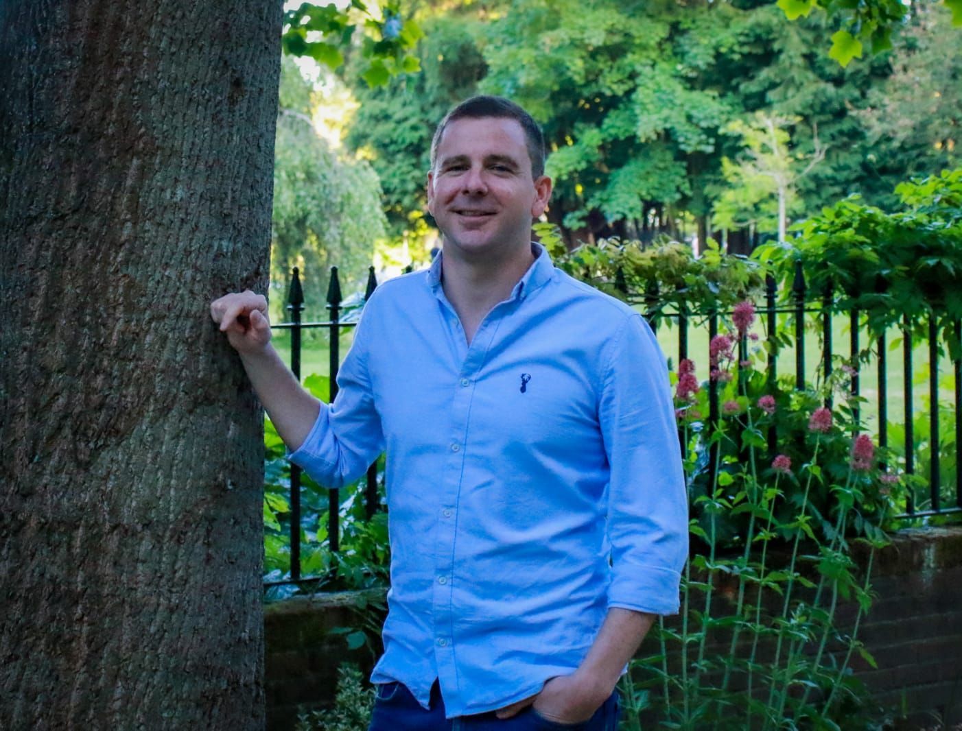 a man in a blue shirt is leaning against a tree in a park .