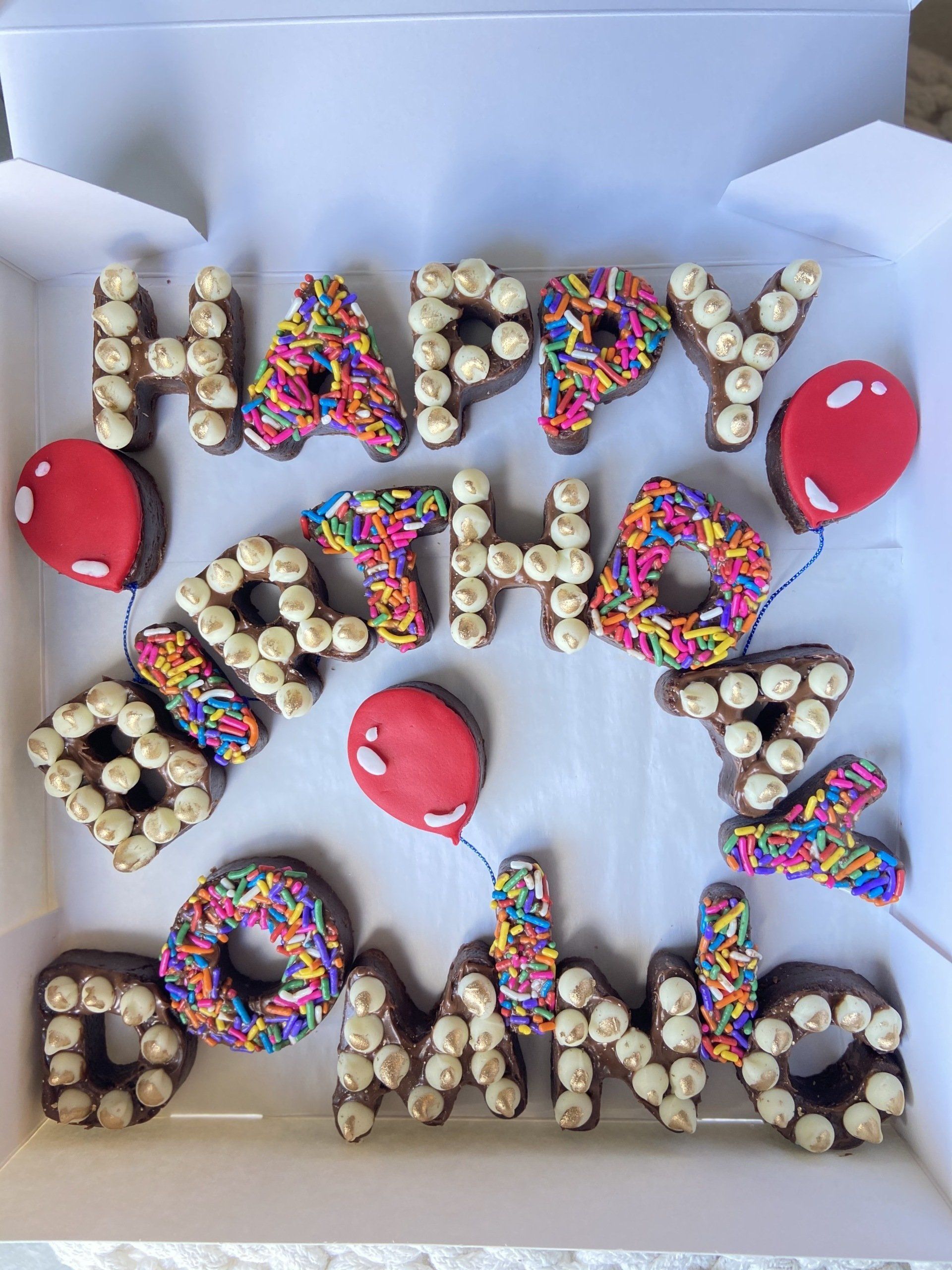 A box of chocolate donuts with sprinkles and balloons that say happy birthday dominic