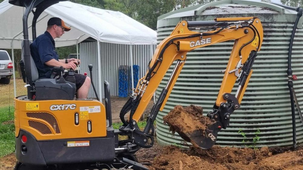A Man Is Driving A Small Excavator Next To A Water Tank — Blue Diesel & Equipment In Speewah, QLD
