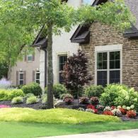 A two-story house with stone and brick exterior sits behind a vibrant landscaped garden featuring shrubs and flowers.
