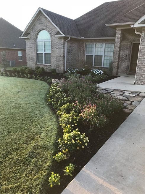 A well-maintained garden bed with various green shrubs and flowers bordering a grassy lawn and a house exterior.