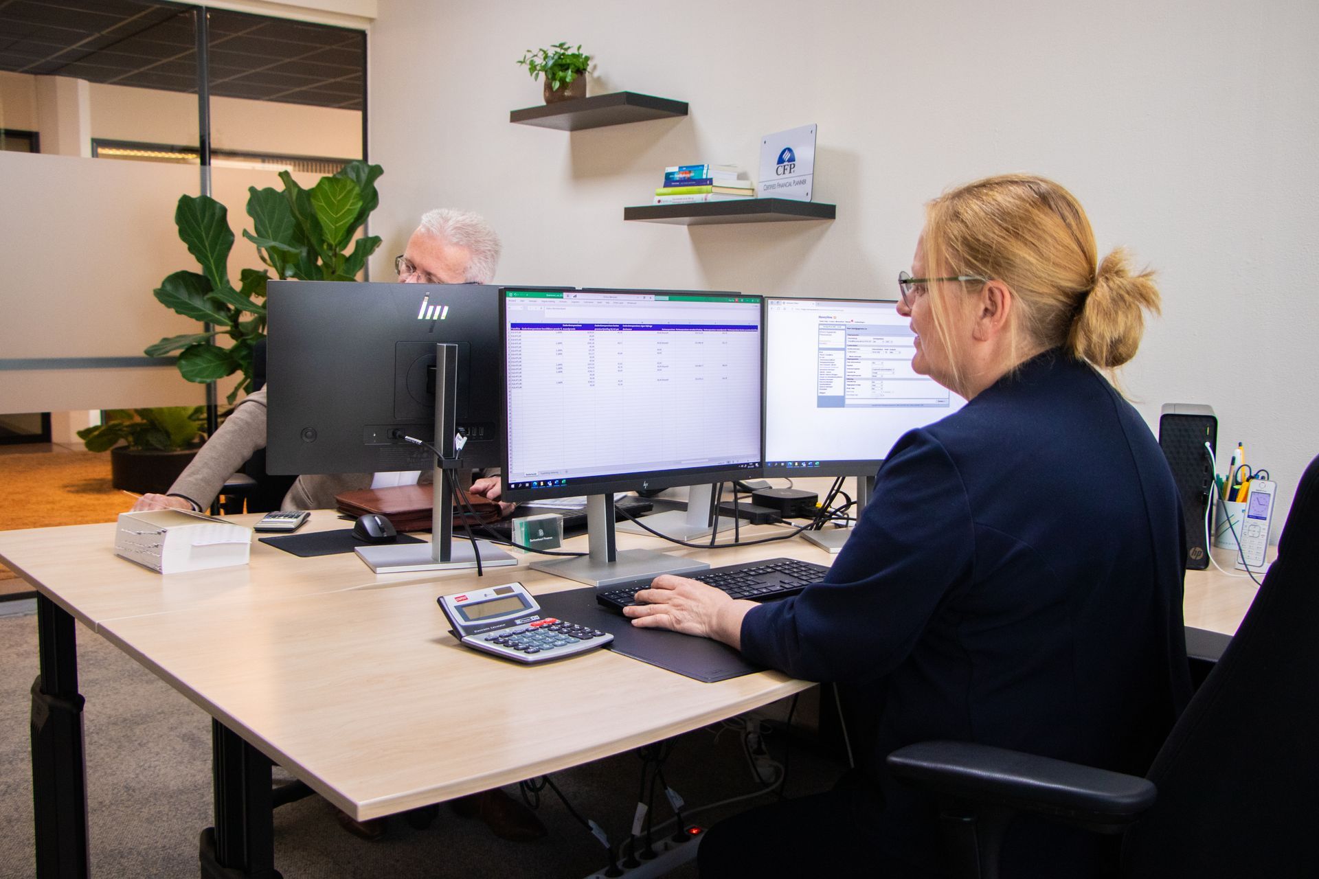 Twee mensen werken achter computers op kantoor. Een vrouw in een blauw jasje typt aan een bureau met twee monitoren.