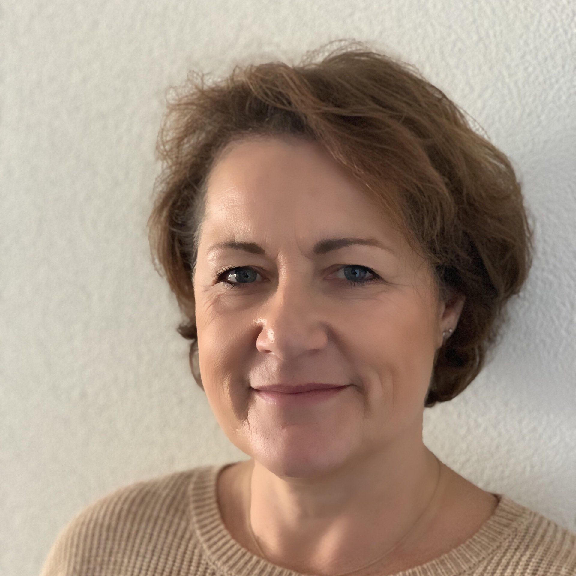 Een vrouw met kort bruin haar staat lachend in een bruine trui tegen een witte muur.