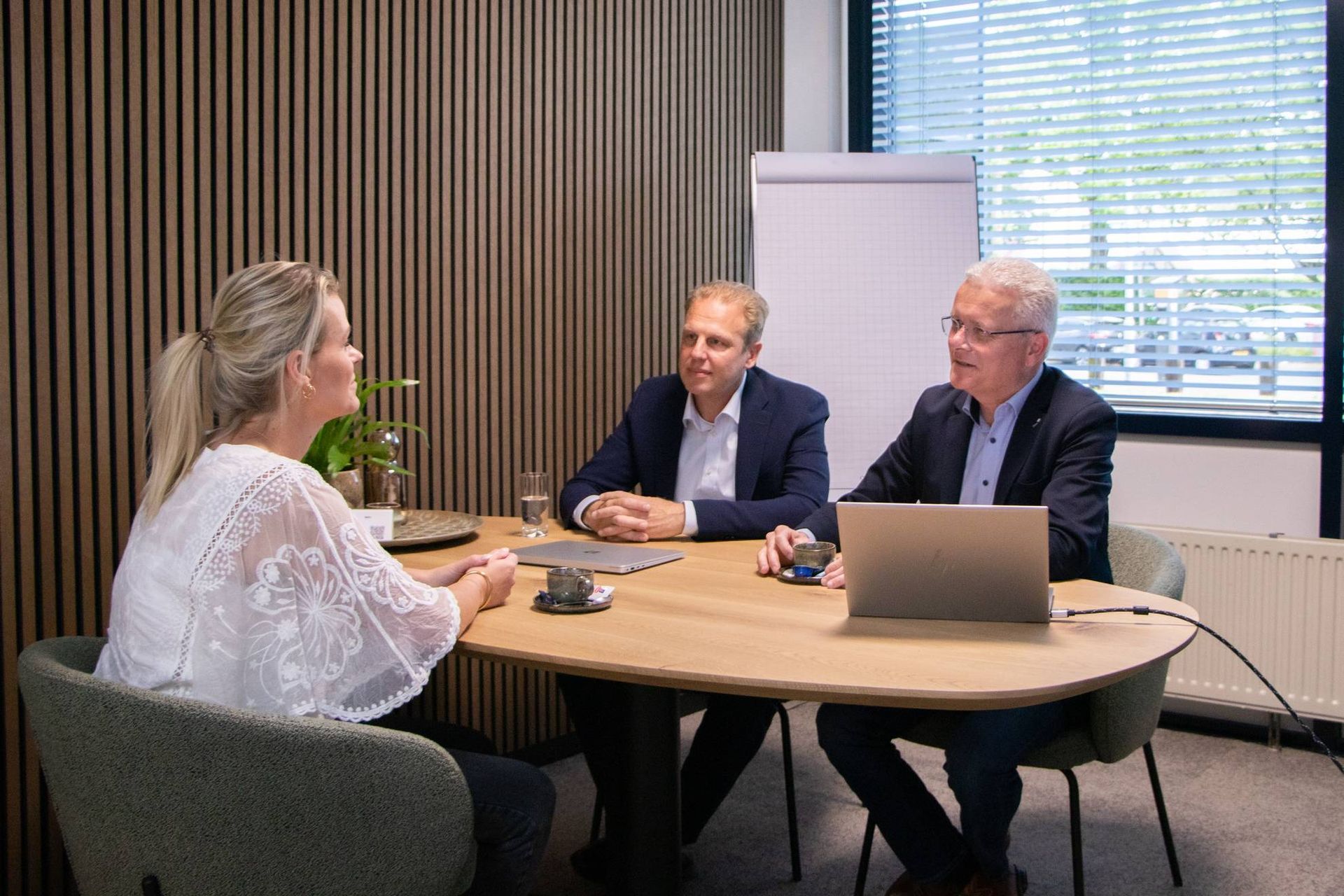 Drie mensen in vergadering op kantoor, discussiëren met elkaar via een laptop en kopjes koffie op een ronde tafel.