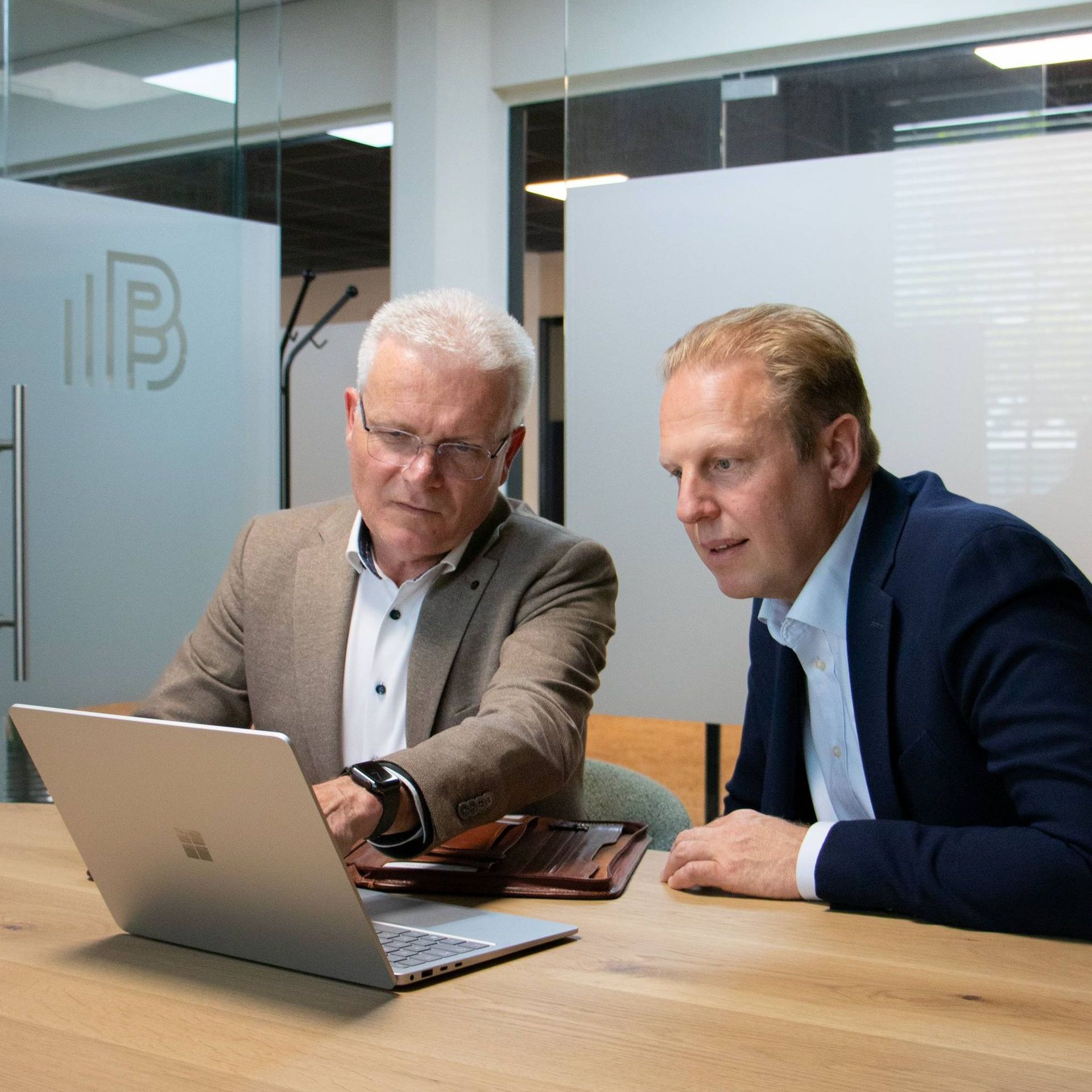Twee mannen op kantoor kijken naar een laptop. De een draagt ​​een bril en een bruin jasje. De ander een blauw pak.
