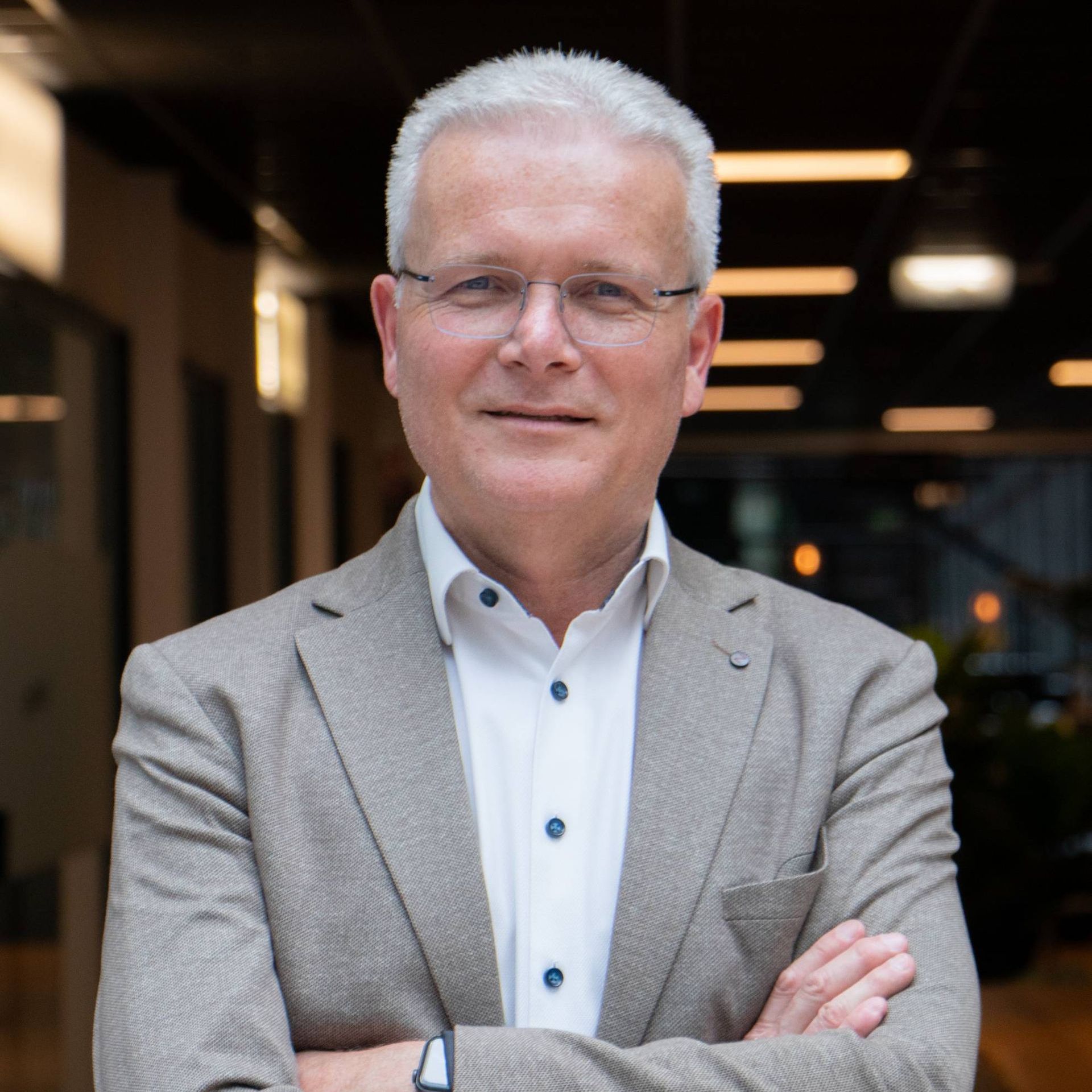 Man met grijs haar en een bril, armen over elkaar, gekleed in een lichtbruin jasje en wit overhemd, binnenshuis.