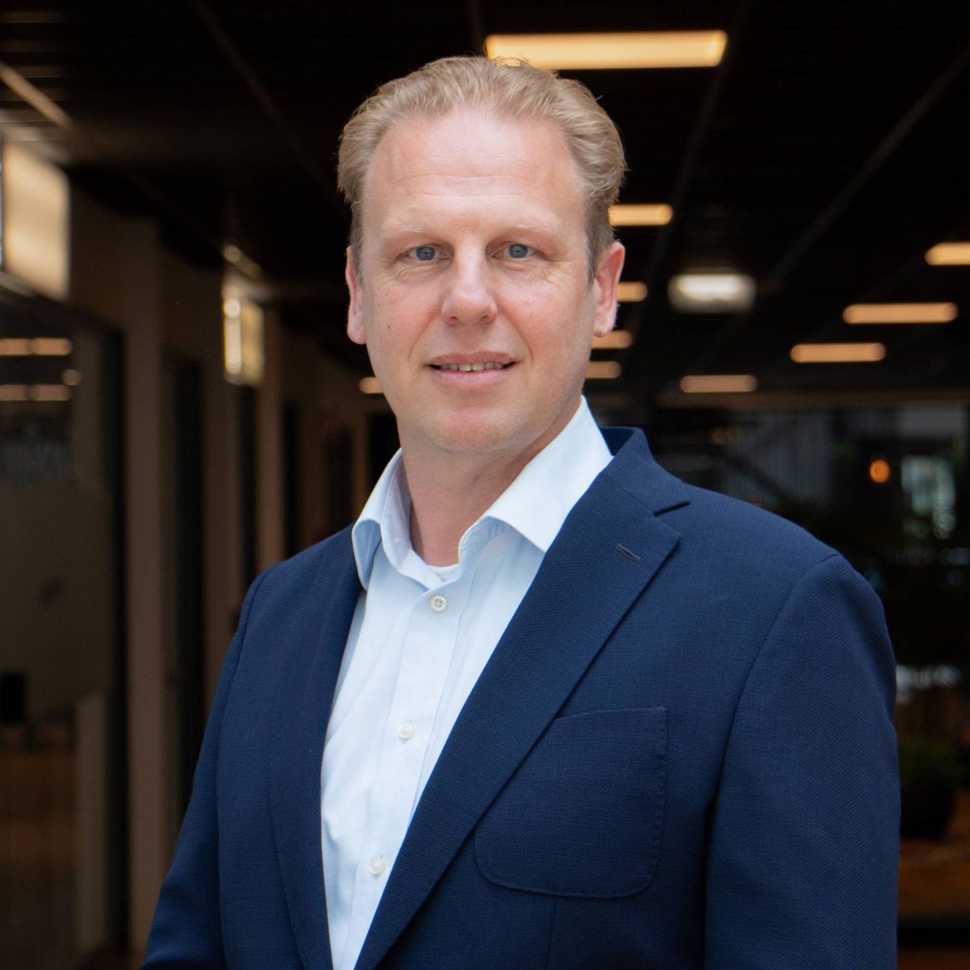 Man in een blauw pak en lichtblauw overhemd lacht in een moderne kantooromgeving.