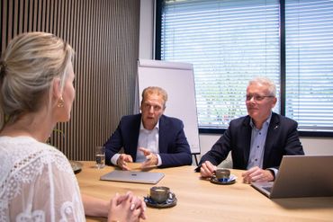 Vrouw in wit kanten topje wordt geïnterviewd door twee mannen in pak aan een tafel.