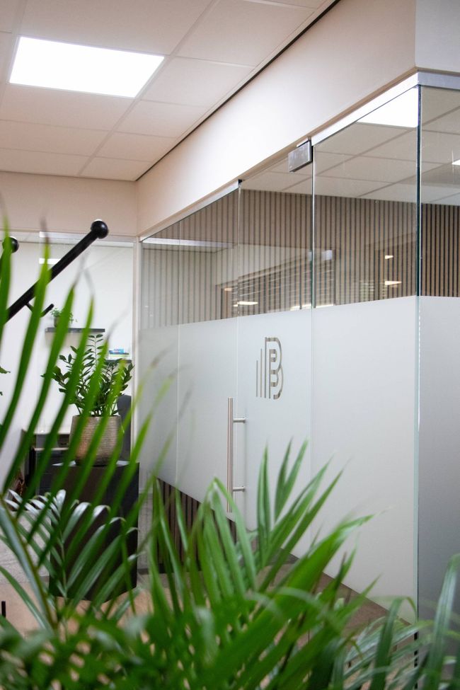 Kantoor met glazen wanden, een matglazen deur en groene planten op de voorgrond; uitzicht van binnen.