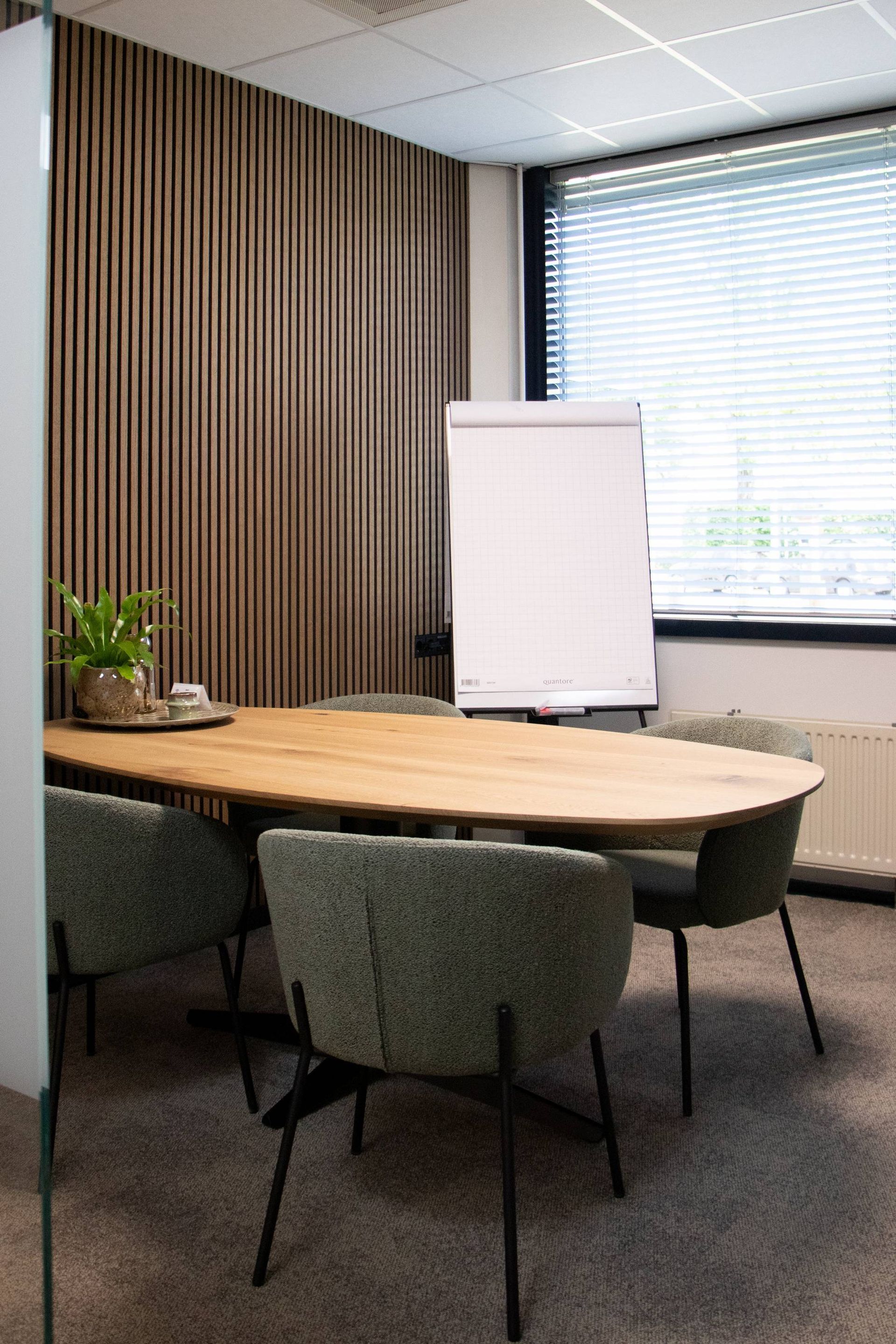 Ovale houten tafel in een vergaderruimte met groene stoelen, whiteboard en houten lambrisering.