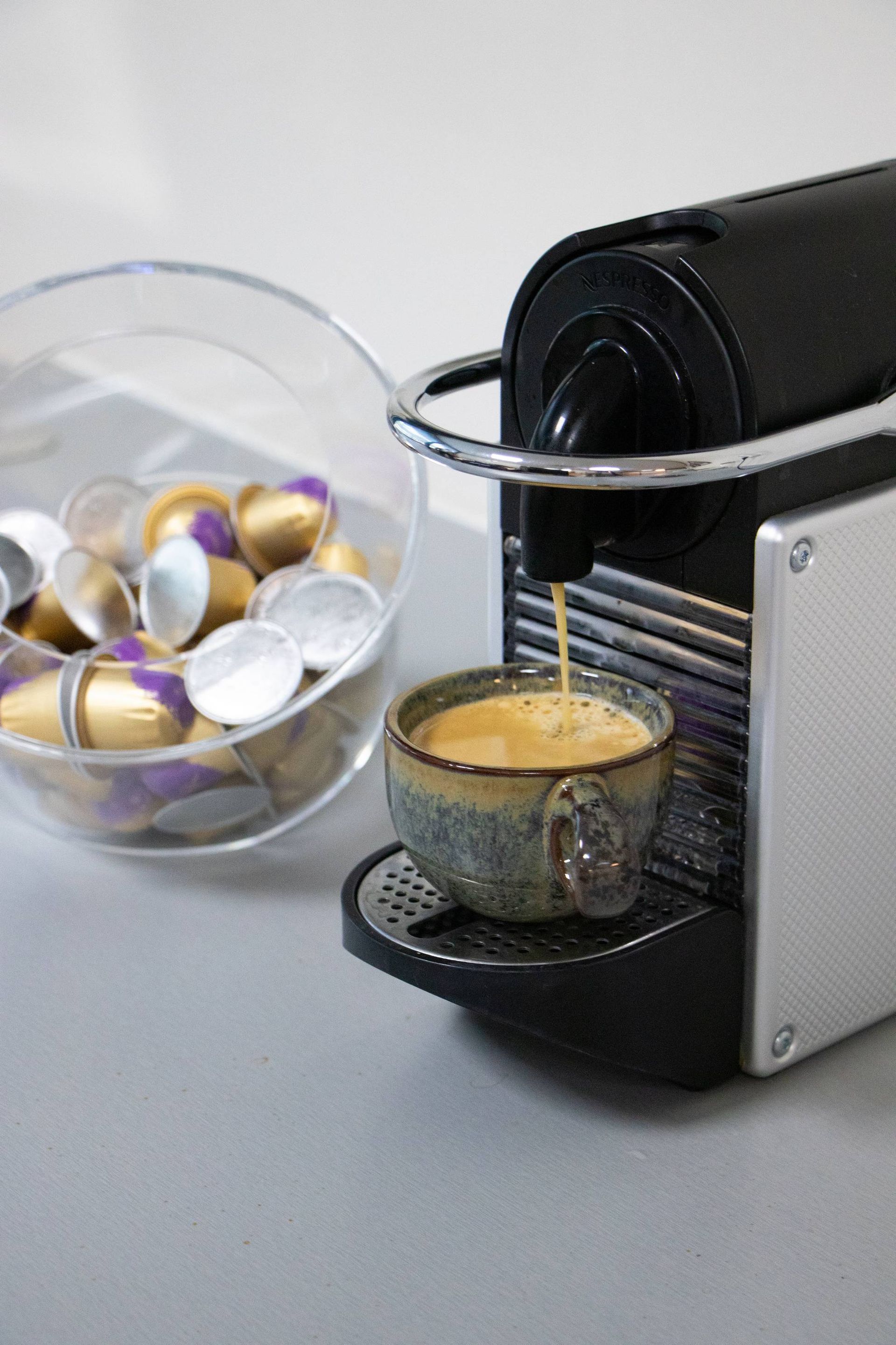Koffiezetapparaat dat espresso in een gebloemd kopje zet; koffiecapsules liggen ernaast in een doorzichtige kom.