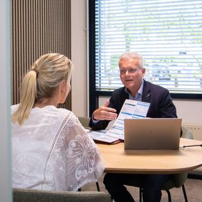 Vrouw in een wit kanten topje is in vergadering met een man die papieren vasthoudt, zittend aan een tafel met een laptop.