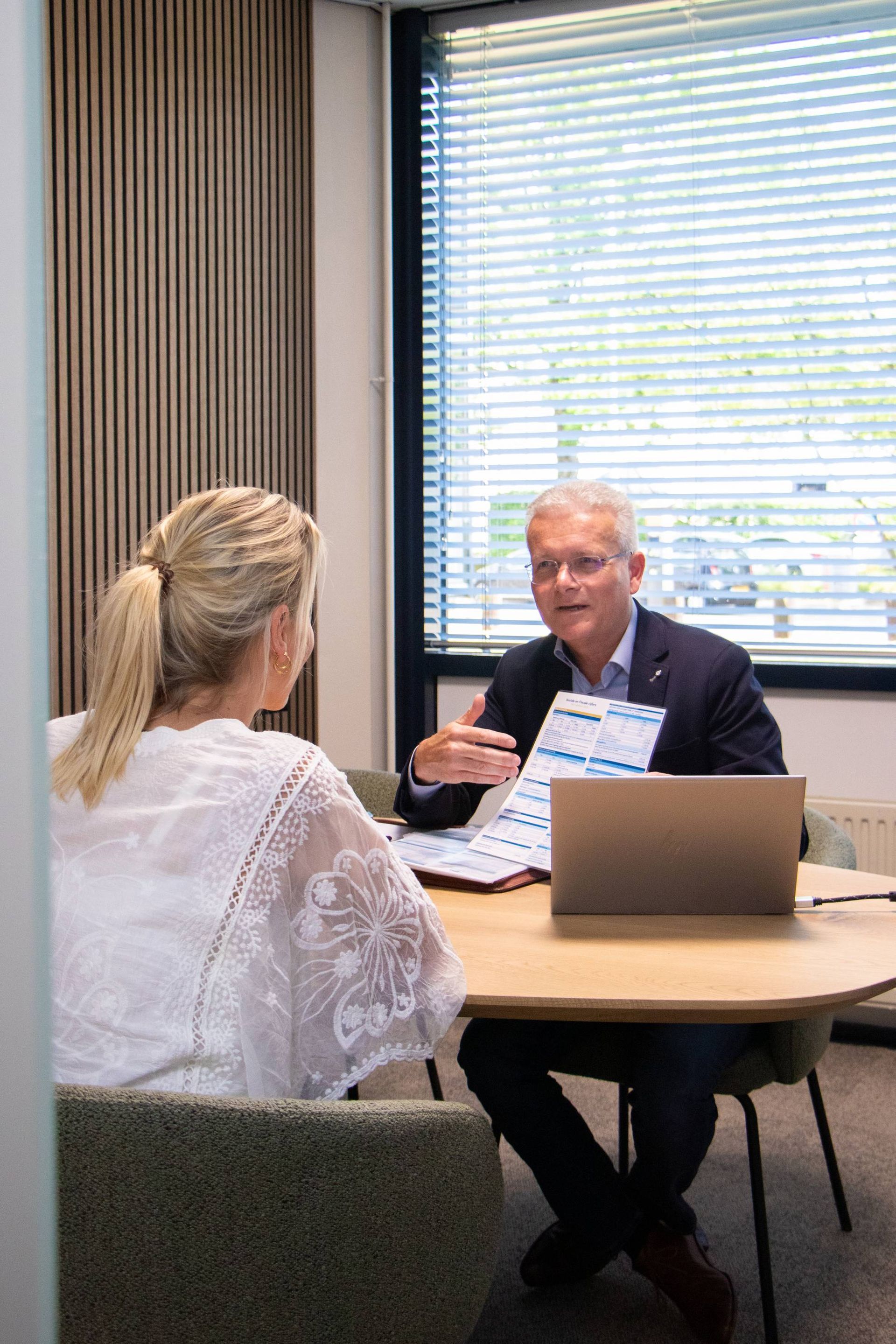 Vrouw in een wit kanten topje is in gesprek met een man in een blazer; ze bespreken documenten aan een tafel.