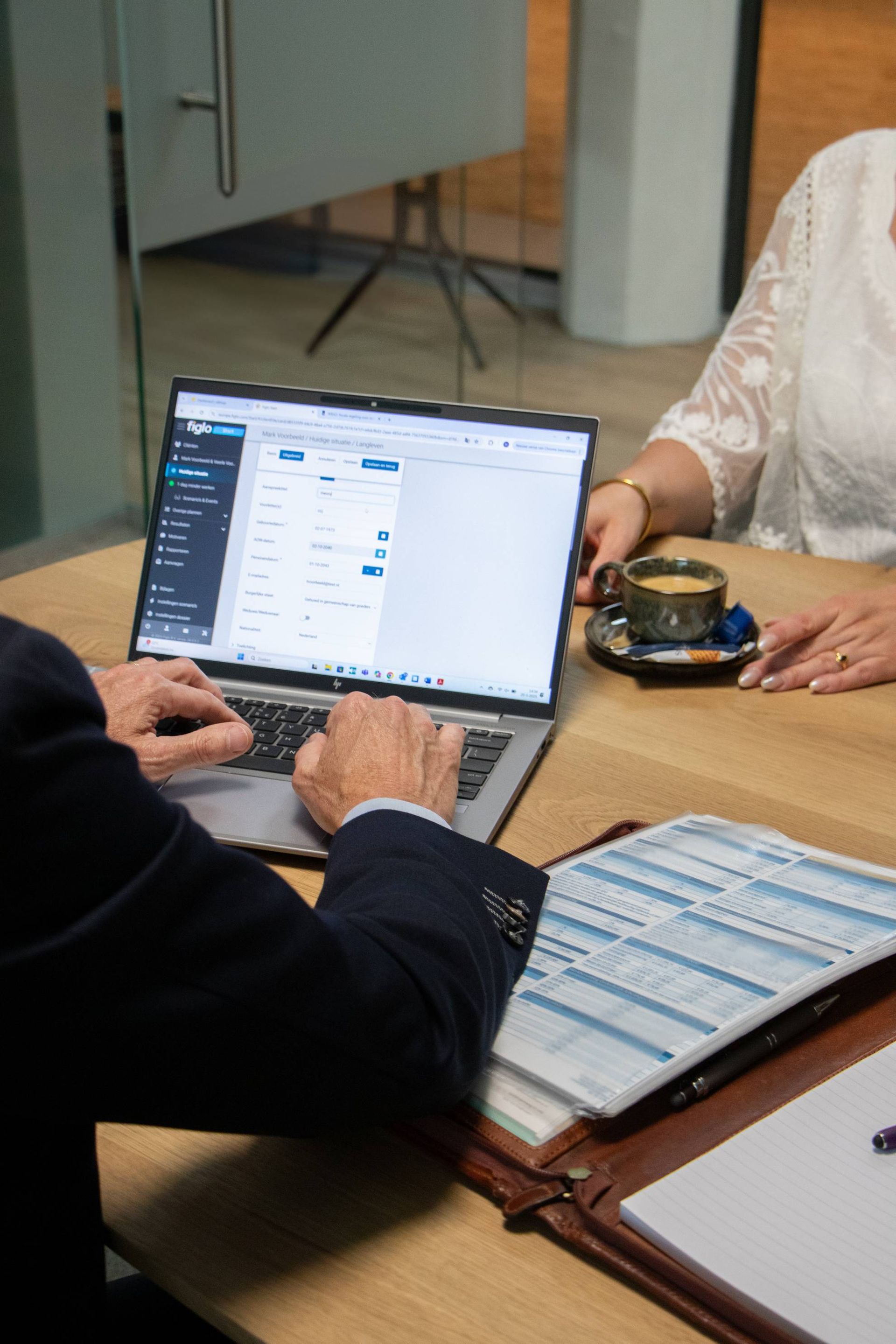 Man en vrouw aan een tafel, man gebruikt laptop, papierwerk voor zich, koffie op tafel.