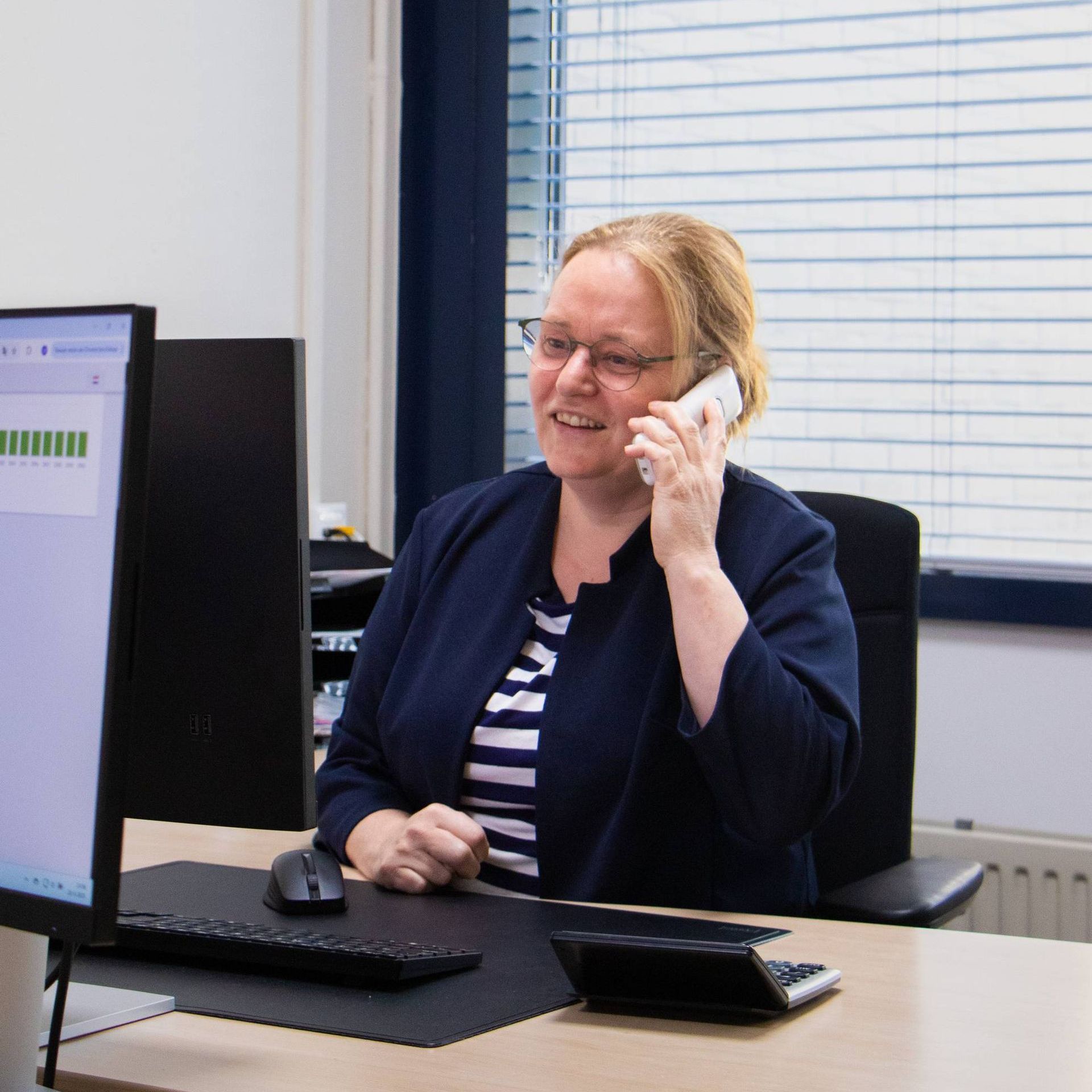 Een vrouw op kantoor praat aan de telefoon en lacht terwijl ze naar het computerscherm kijkt.