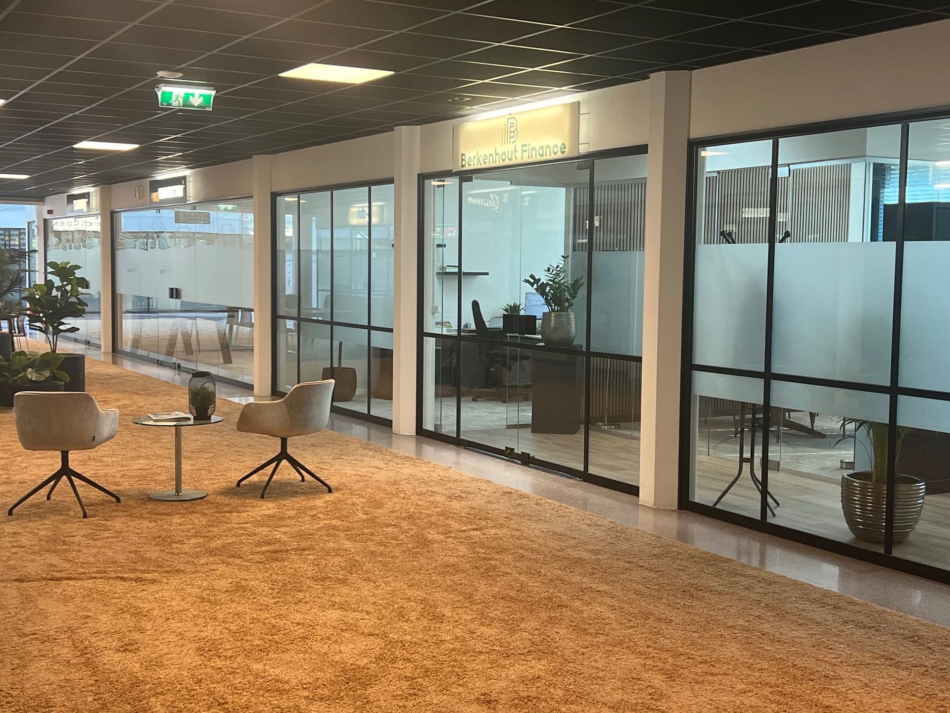 Een kantoorgang met kamers met glazen wanden, beige tapijt en twee stoelen tegenover een klein tafeltje.